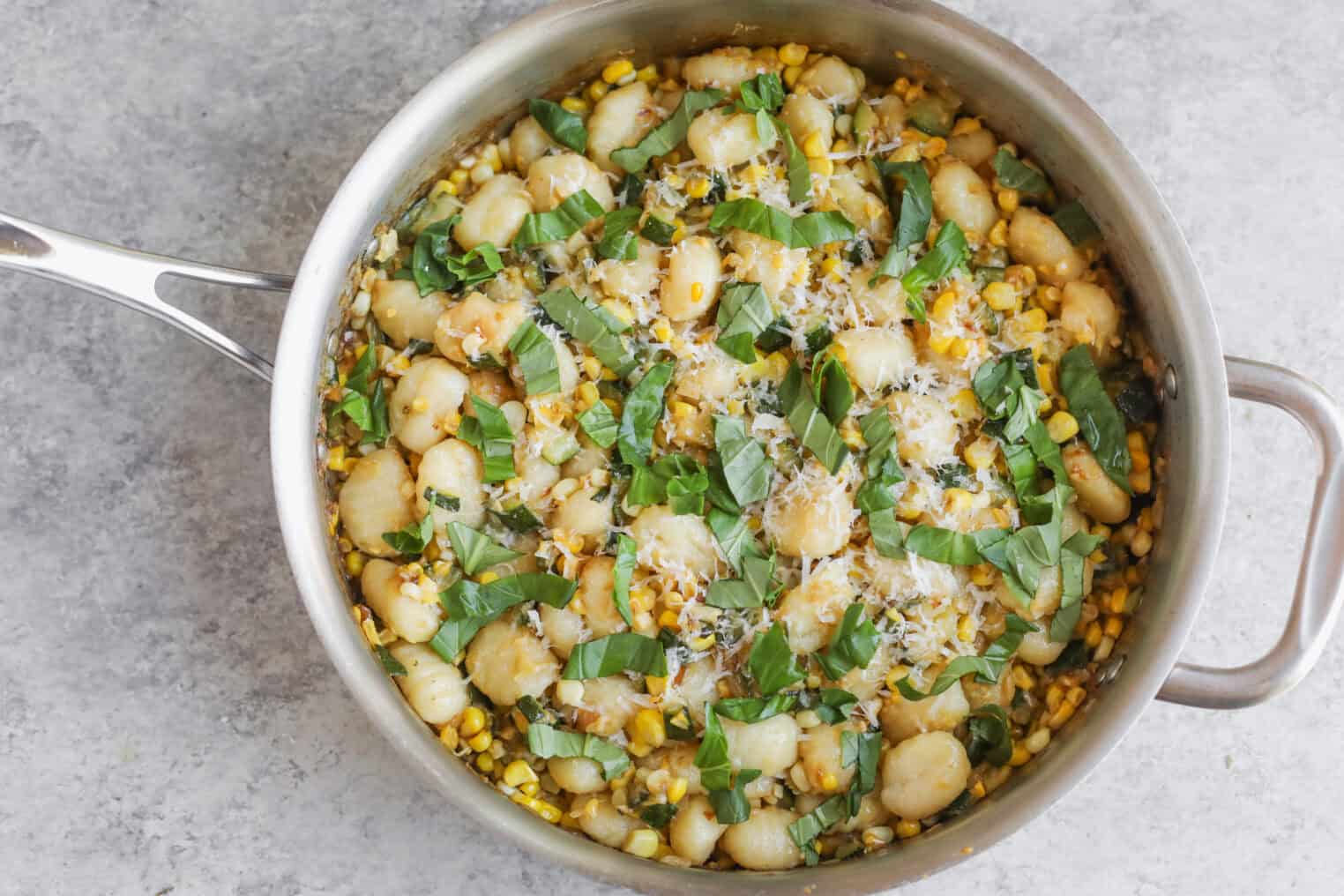 A Pan Filled With Summer Gnocchi, Fresh Corn, And Basil Leaves, Topped With Grated Cheese, Sits On A Light Gray Surface.