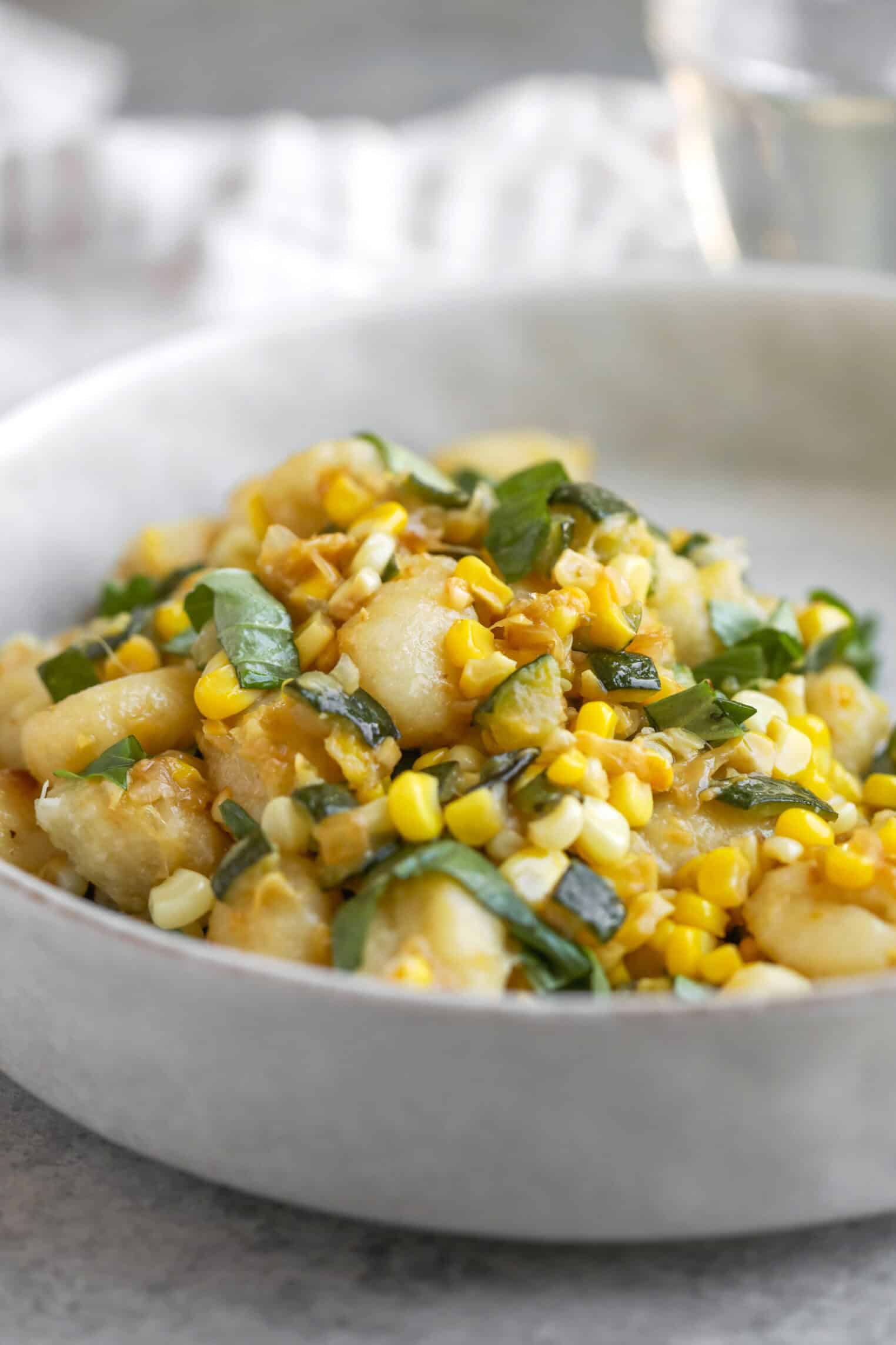 A Bowl Of Summer Gnocchi Topped With Sautéed Fresh Corn, Chopped Basil, And Zucchini, Served In A Light-Colored Dish With A Blurred Background.