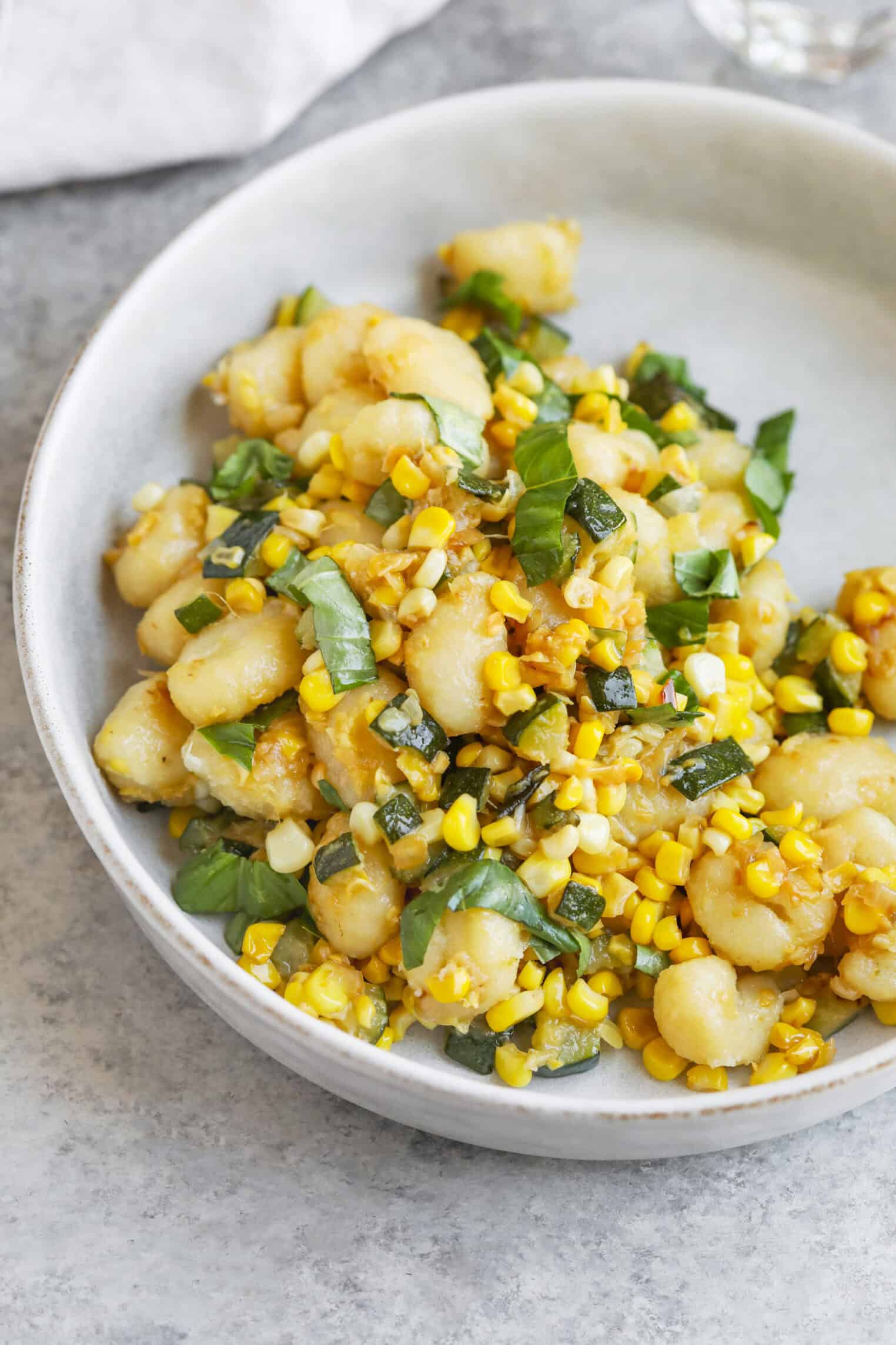 A White Bowl Filled With Summer Gnocchi, Fresh Corn Kernels, And Basil, All Tossed Together On A Light Gray Surface.