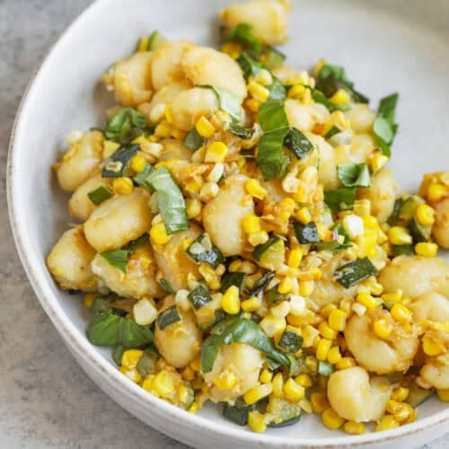 A White Bowl Filled With Summer Gnocchi, Fresh Corn Kernels, And Basil, All Tossed Together On A Light Gray Surface.