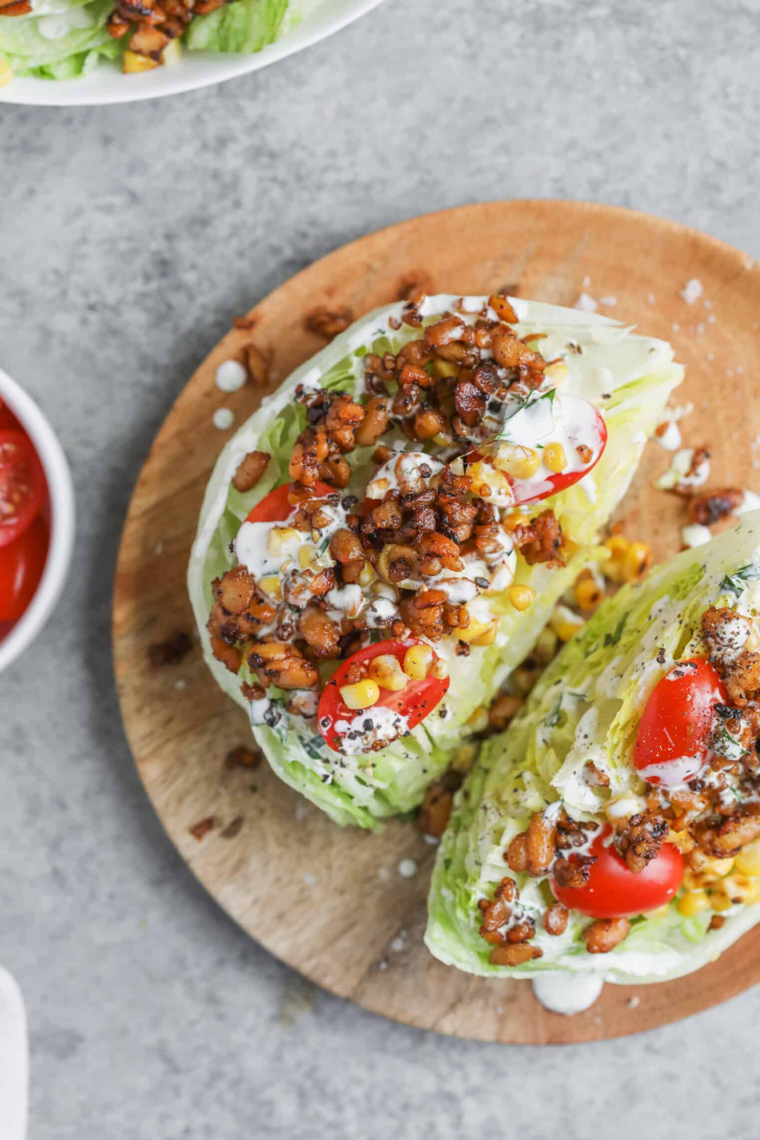 Two Summer Wedge Salads On A Wooden Plate, Topped With Cherry Tomatoes, Corn, Crispy Chickpeas, Herbs, And Drizzled With Creamy Dressing. A Bowl Of Tomatoes Is Partially Visible Beside This Vibrant Summer Dish.