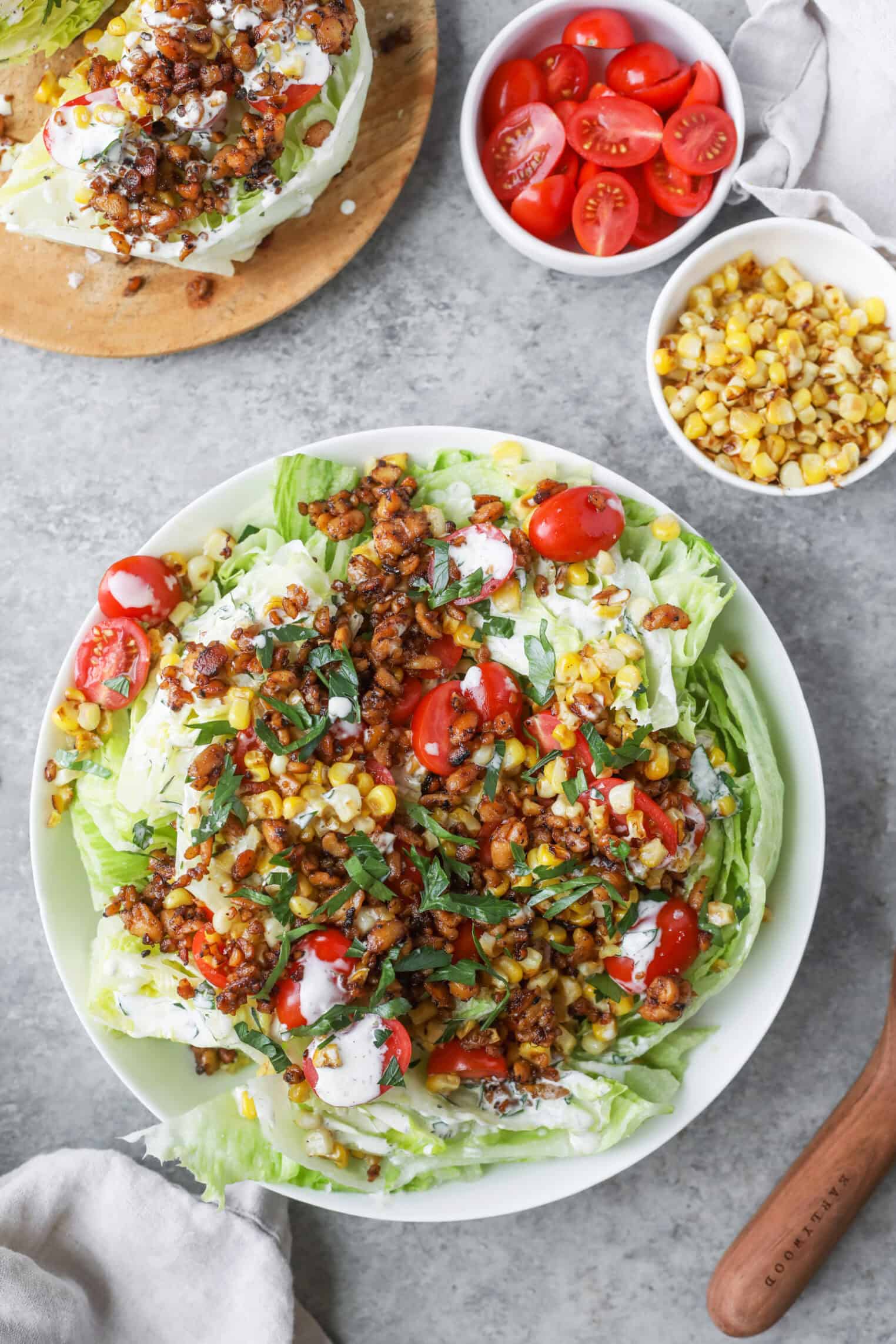 A Plate Of Summer Wedge Salad Topped With Cherry Tomatoes, Corn, Crumbled Tempeh, Fresh Herbs, And A Drizzle Of Creamy Dressing Sits On A Gray Surface. Nearby Are Bowls Of Cherry Tomatoes And Corn, And Another Salad.