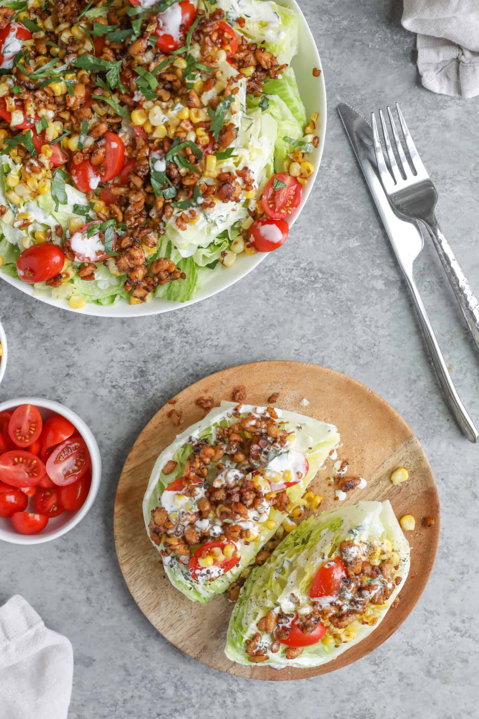 Two Summer Wedge Salads Topped With Chopped Tomatoes, Corn, Ground Meat, Fresh Herbs, And Creamy Dressing Are Served On A Round Wooden Board And A White Plate, With A Small Bowl Of Cherry Tomatoes And Utensils Nearby.