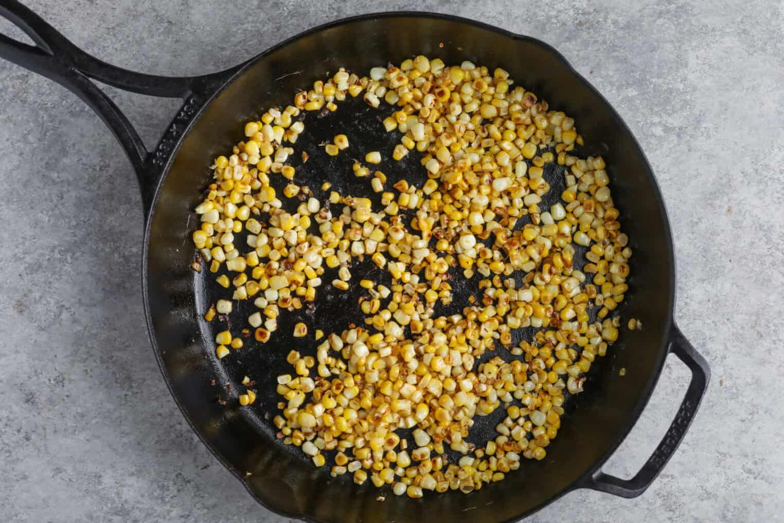 Charred Corn Kernels Are Scattered In A Large Cast Iron Skillet On A Grey, Textured Surface—The Perfect Topping For A Summer Wedge Salad. The Corn Is Golden Brown, Indicating It Has Been Sautéed Or Roasted.