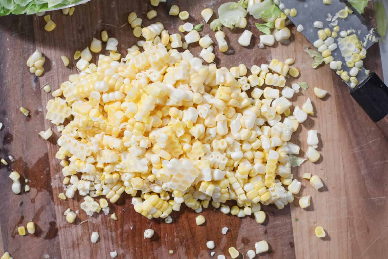 A Pile Of Freshly Cut Corn Kernels Sits On A Wooden Cutting Board Next To A Large Kitchen Knife, Ready To Be Added To A Summer Wedge Salad. Scattered Corn Pieces And Leaves Add A Rustic Touch.