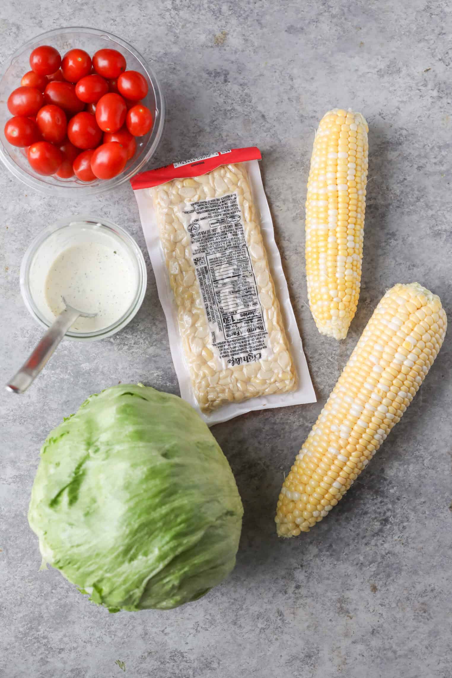 A Head Of Iceberg Lettuce, Two Ears Of Corn, A Block Of Tempeh In Packaging, Cherry Tomatoes, And Creamy Dressing Are Arranged On A Gray Surface—Perfect Ingredients For A Fresh Summer Wedge Salad.