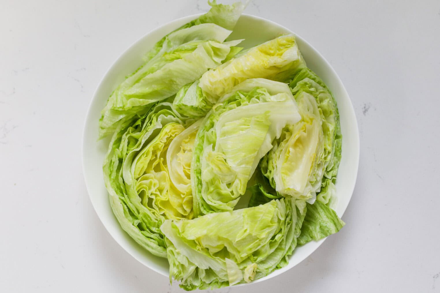 A White Bowl Filled With Several Wedges Of Fresh Iceberg Lettuce, Perfect For A Summer Wedge Salad, Sits On A White Surface. The Pale Green Lettuce Boasts Crisp, Layered Leaves.