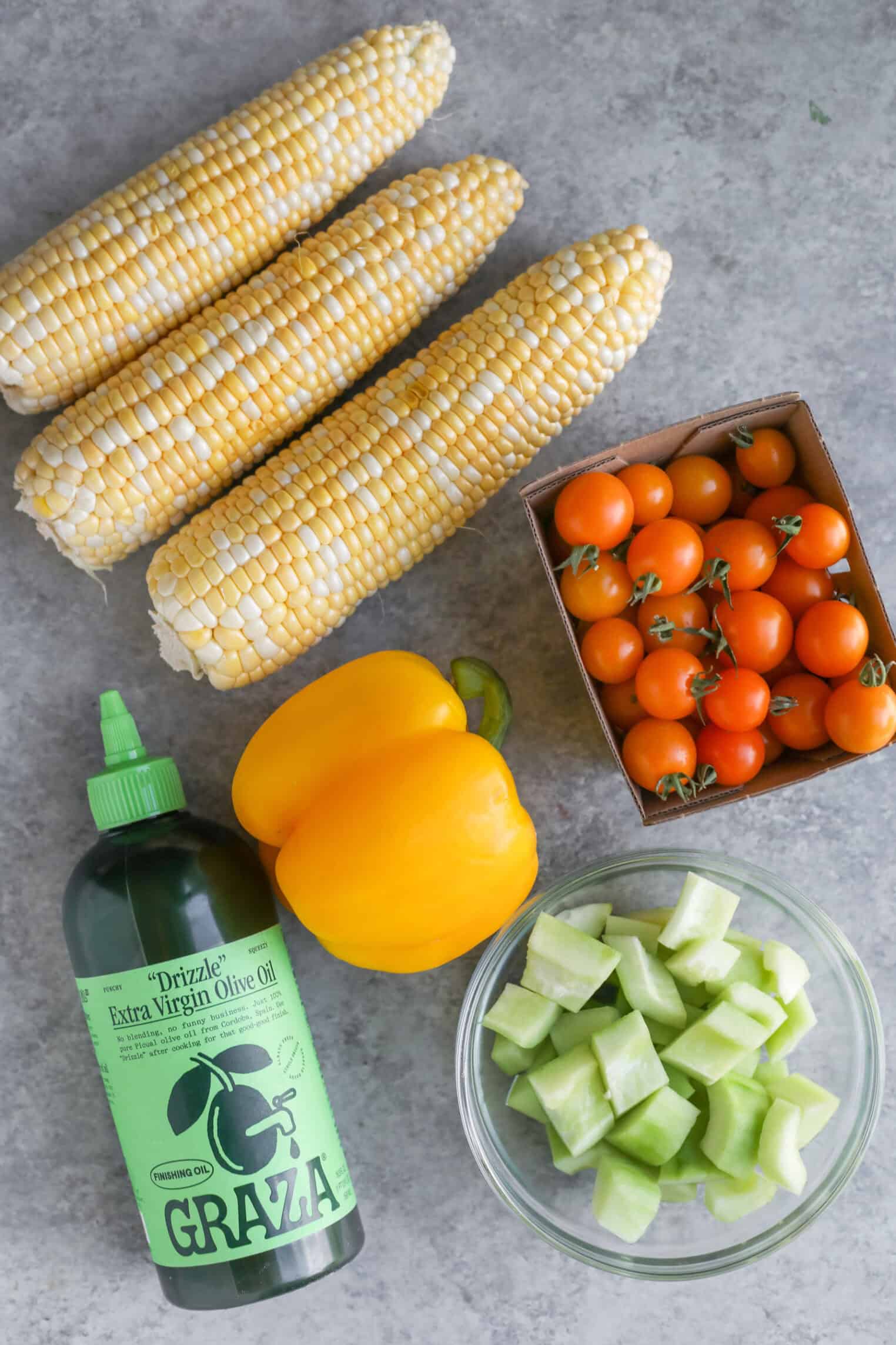 Sun Gold Tomato and Corn Gazpacho 2 Three Ears Of Corn, A Box Of Sun Gold Tomatoes, A Yellow Bell Pepper, Sliced Cucumbers, And Graza Extra Virgin Olive Oil Arranged On A Gray Surface—Perfect Ingredients For A Fresh Corn Gazpacho.