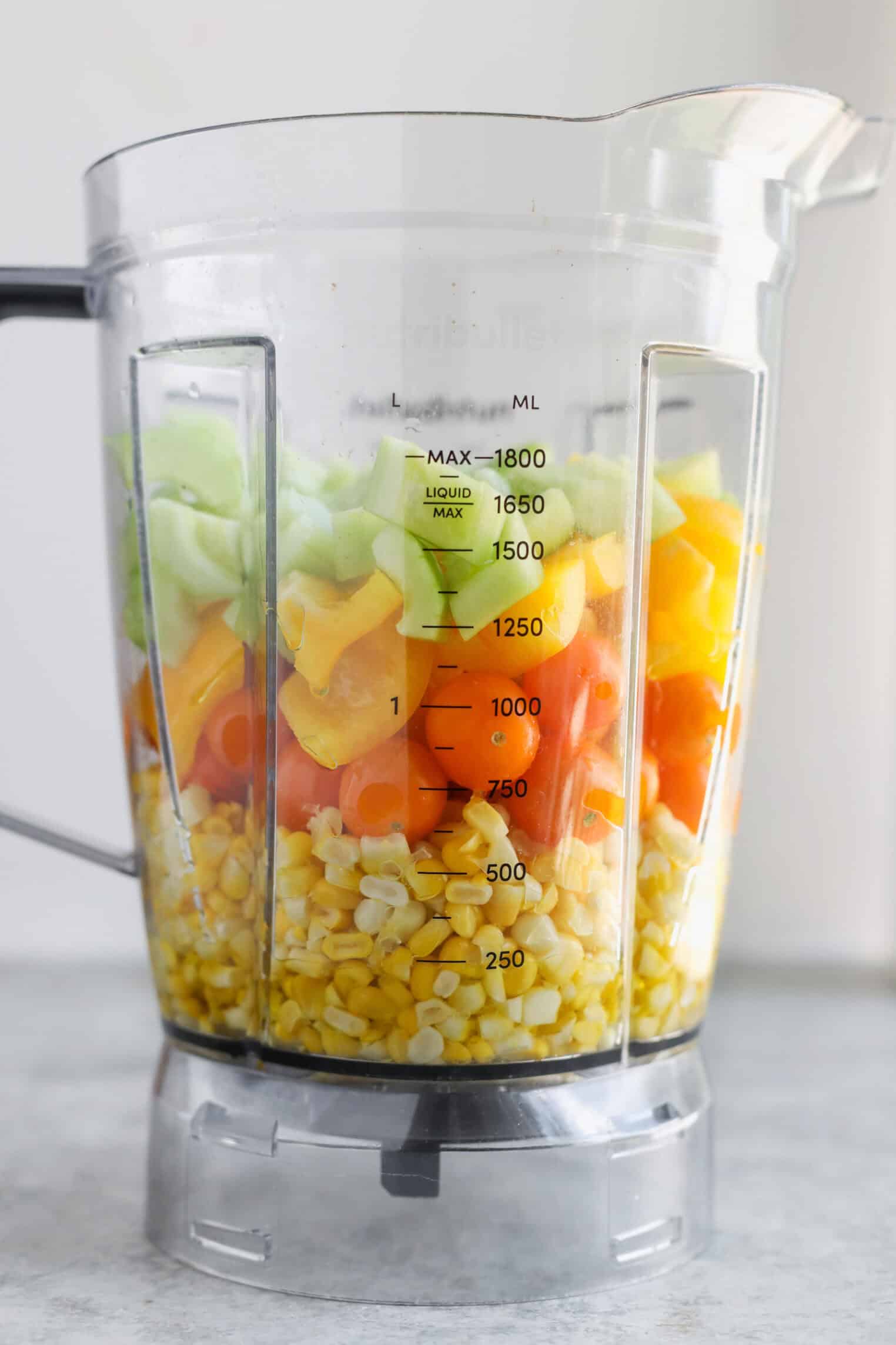 Sun Gold Tomato and Corn Gazpacho 3 A Blender Container Filled With Layers Of Fresh Yellow Corn Kernels, Orange Carrots, Yellow Bell Peppers, And Green Celery Sits On A Gray Countertop—Perfect For Preparing A Vibrant Corn Gazpacho.