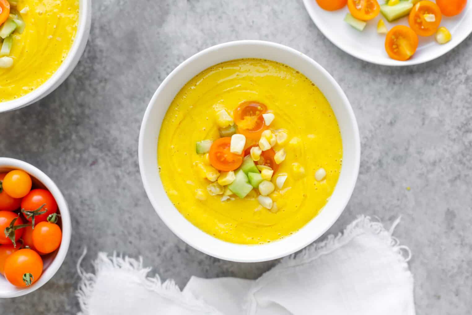 Sun Gold Tomato and Corn Gazpacho 5 A Bowl Of Creamy Yellow Corn Gazpacho Garnished With Halved Cherry Tomatoes, Diced Cucumber, And Corn Kernels, Placed On A Gray Surface Next To Fresh Sun Gold Tomatoes And A White Napkin.