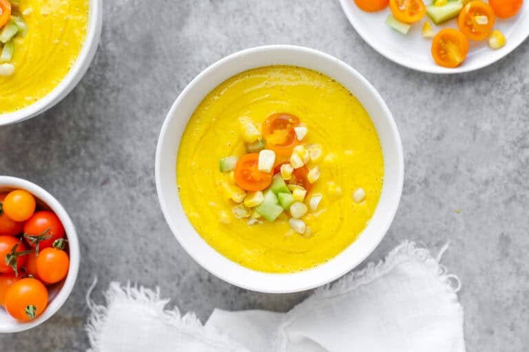 A Bowl Of Creamy Yellow Corn Gazpacho Garnished With Halved Cherry Tomatoes, Diced Cucumber, And Corn Kernels, Placed On A Gray Surface Next To Fresh Sun Gold Tomatoes And A White Napkin.