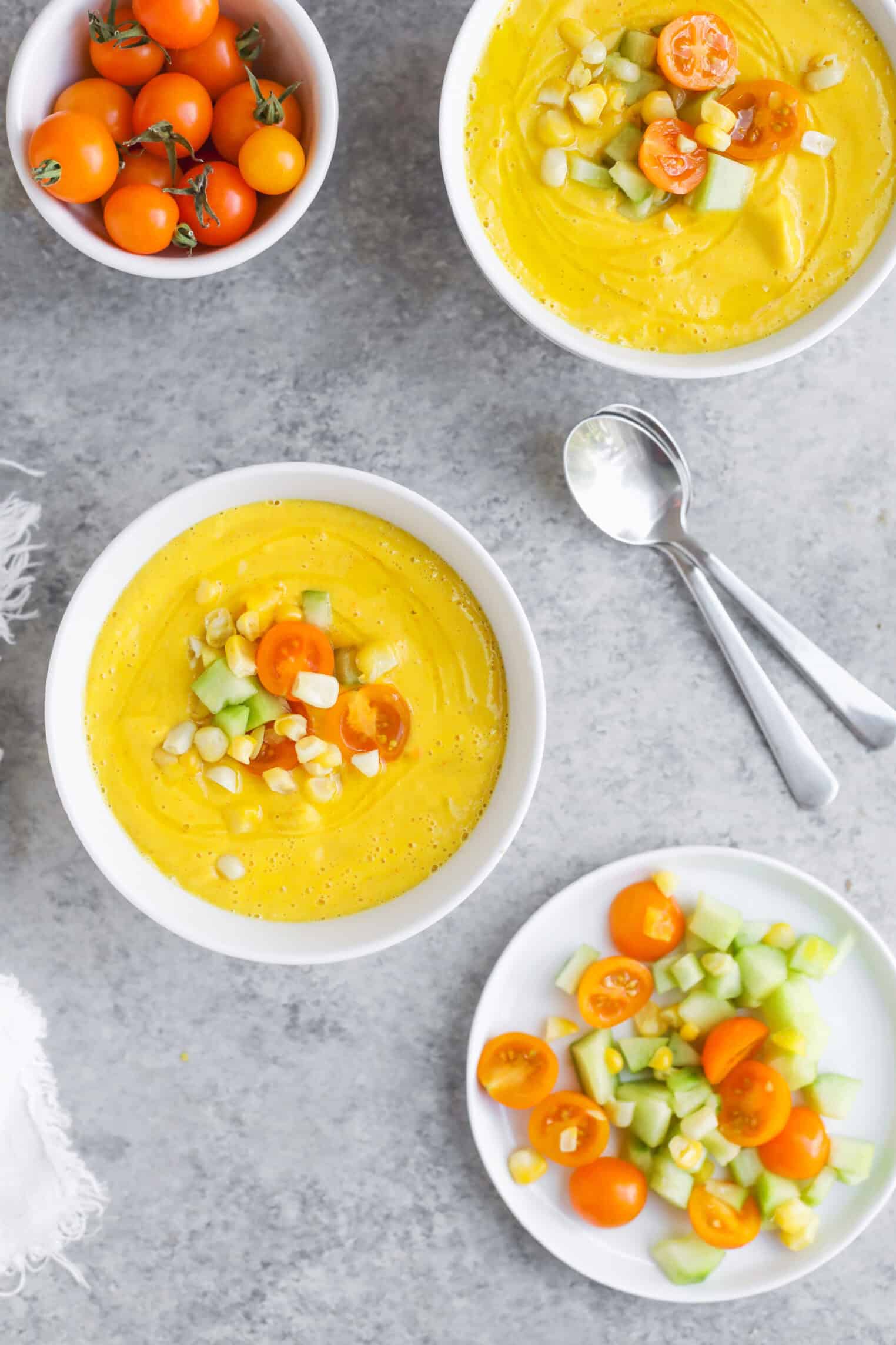 Sun Gold Tomato and Corn Gazpacho 6 Three Bowls Of Creamy Yellow Corn Gazpacho Topped With Diced Cucumber, Corn, And Halved Sun Gold Tomato, Alongside A Plate Of Toppings, A Bowl Of Cherry Tomatoes, And Two Spoons On A Gray Surface.