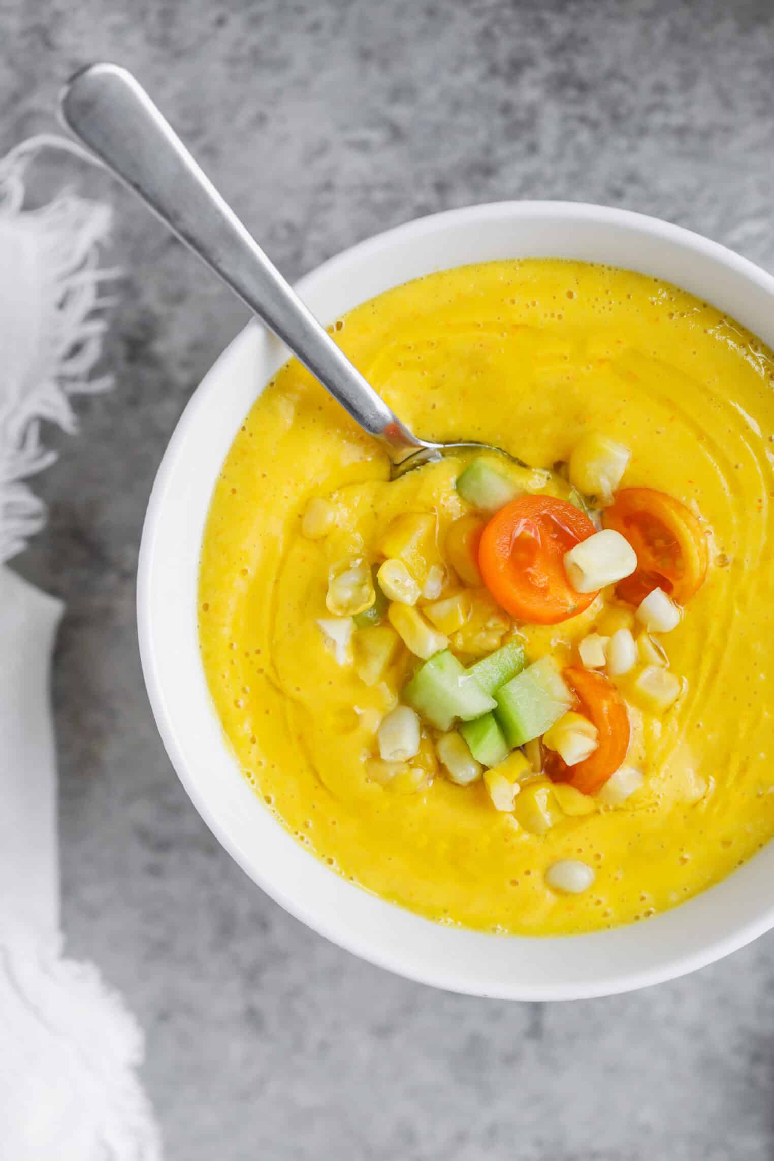 Sun Gold Tomato and Corn Gazpacho 1 A Bowl Of Creamy Yellow Corn Gazpacho Topped With Diced Cucumber, Corn Kernels, And Halved Cherry Tomatoes, With A Spoon Resting Inside The Bowl On A Gray Surface.