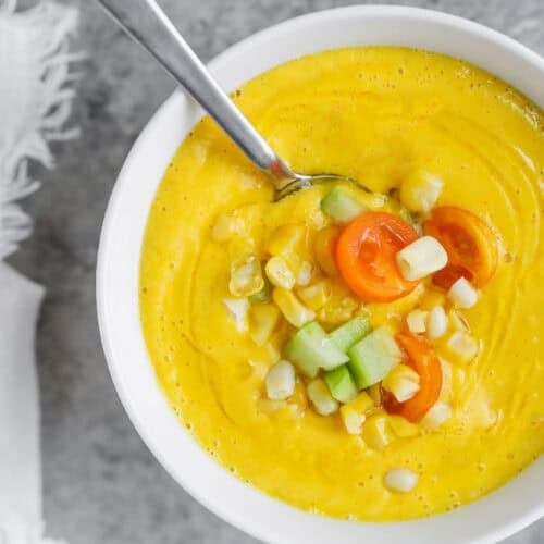 Sun Gold Tomato and Corn Gazpacho 9 A Bowl Of Creamy Yellow Corn Gazpacho Topped With Diced Cucumber, Corn Kernels, And Halved Cherry Tomatoes, With A Spoon Resting Inside The Bowl On A Gray Surface.