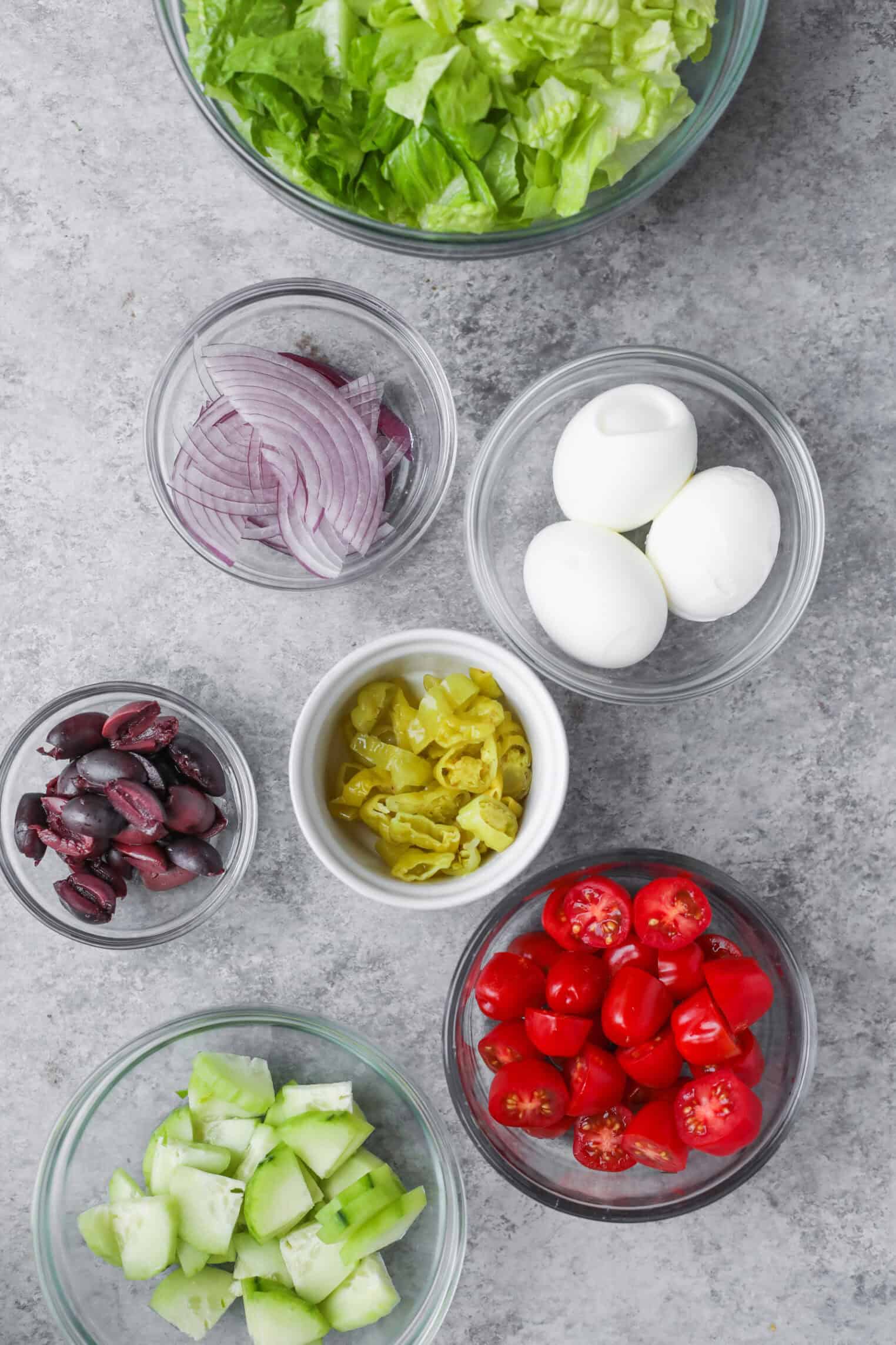 Seven Glass Bowls On A Gray Surface, Each Featuring Ingredients For A Tarpon Springs Greek Salad With Potato Salad: Chopped Lettuce, Sliced Red Onion, Whole Hard-Boiled Eggs, Black Olives, Pepperoncini, Cherry Tomatoes, And Cucumber.