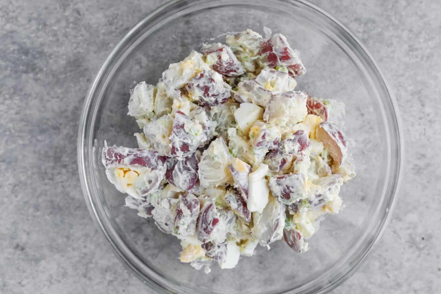 A Glass Bowl Filled With Creamy Potato Salad—Reminiscent Of Tarpon Springs Greek Salad With Potato Salad—Made With Red Potatoes, Chopped Hard-Boiled Eggs, Celery, And Green Onions, Set On A Gray Countertop.