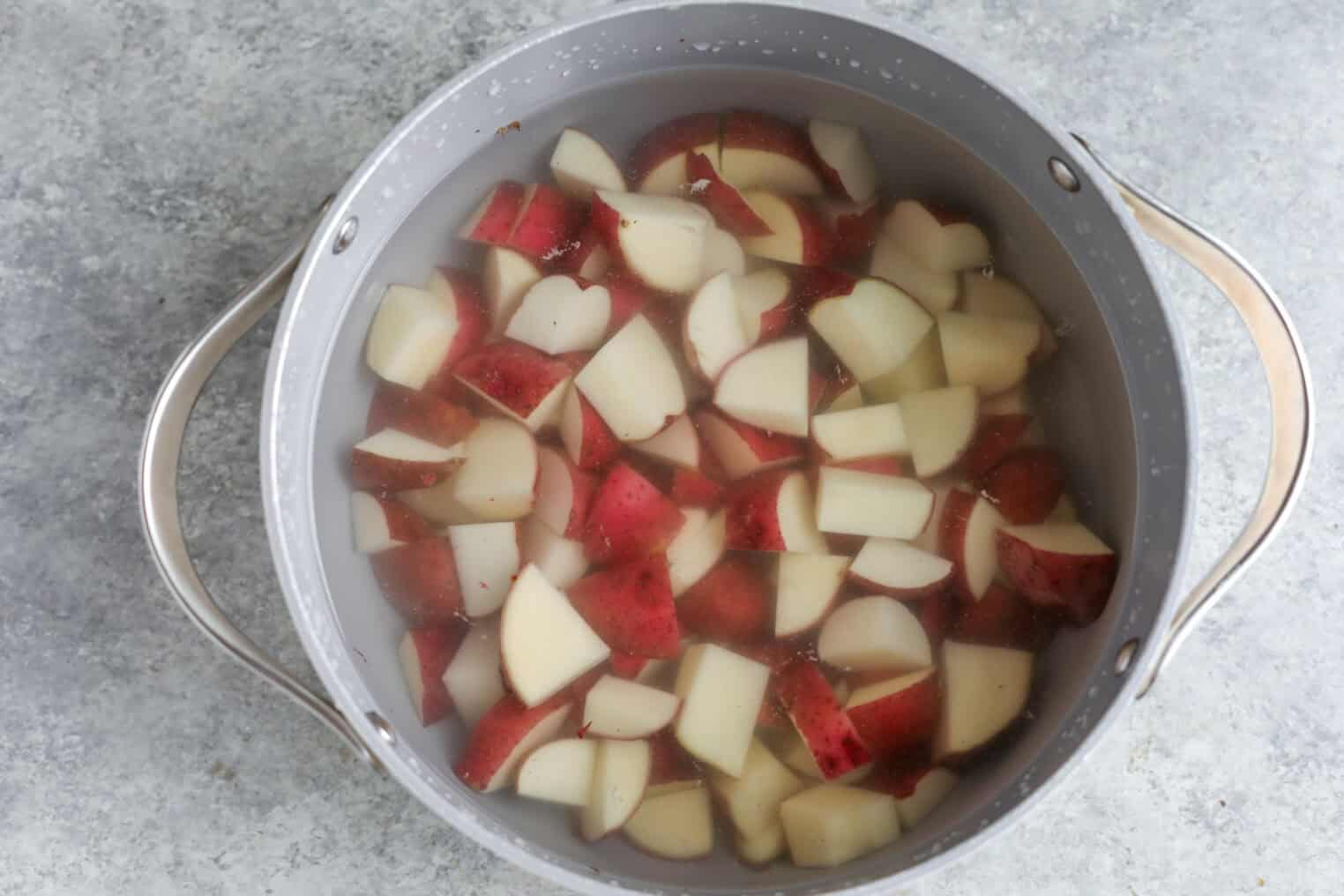 A Pot Filled With Water And Chopped Red-Skinned Potatoes, Some With Skin On, Soaking Before Cooking—Just Like The Potatoes Used In A Classic Tarpon Springs Greek Salad With Potato Salad. The Pot Sits On A Gray Countertop, Ready For The Next Step.