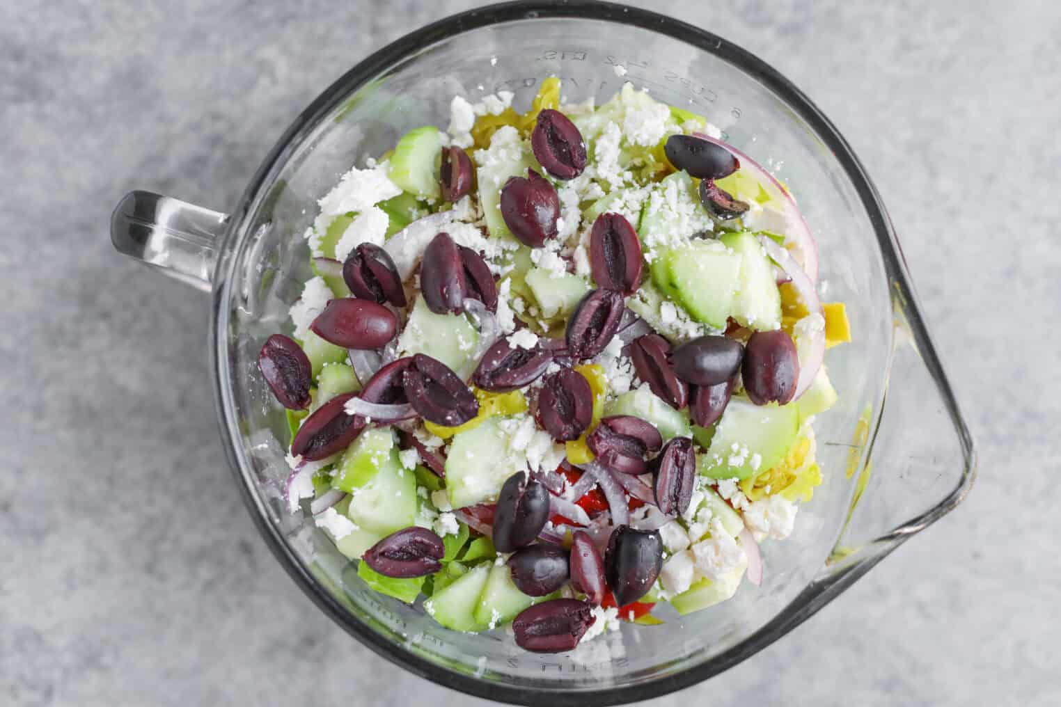 A Glass Bowl Filled With Tarpon Springs Greek Salad With Potato Salad, Featuring Sliced Cucumbers, Red Onions, Green Bell Peppers, Feta Cheese, And Kalamata Olives, Viewed From Above On A Gray Surface.