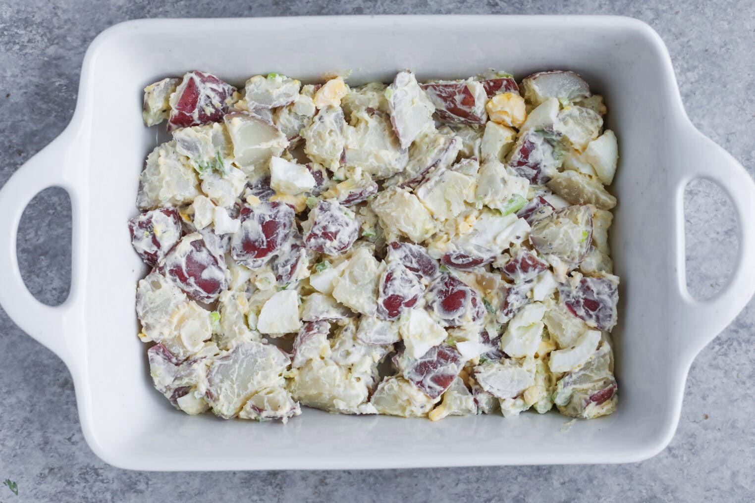 A White Baking Dish Filled With Creamy Potato Salad Inspired By Tarpon Springs Greek Salad With Potato Salad, Made From Red Potatoes, Chopped Eggs, And Green Herbs, Placed On A Gray Countertop.