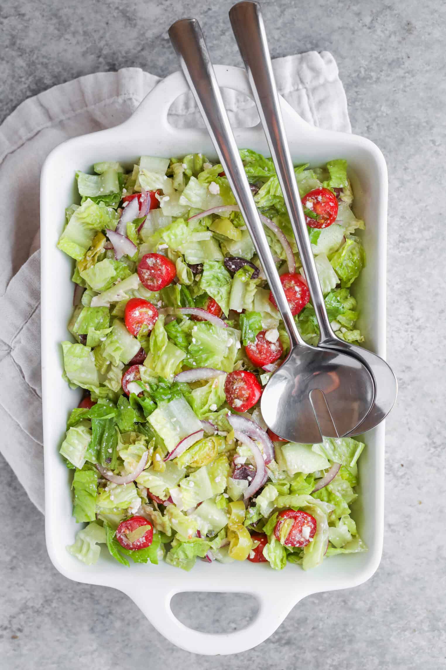 A Rectangular White Dish Filled With A Fresh Tarpon Springs Greek Salad With Potato Salad, Featuring Lettuce, Cherry Tomatoes, Red Onion, And Olives. Two Metal Serving Utensils Rest On Top, Set On A Light-Colored Napkin Over A Grey Surface.
