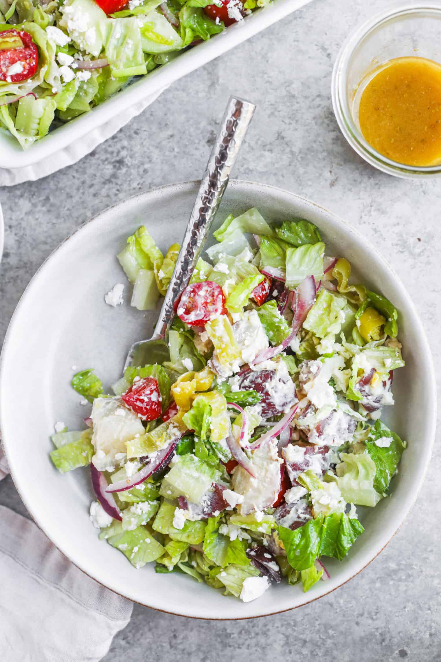 A Bowl Of Fresh Tarpon Springs Greek Salad With Potato Salad, Lettuce, Tomatoes, Cucumber, Red Onion, Feta Cheese, And Dressing On The Side, Served With A Fork On A Light Gray Surface.