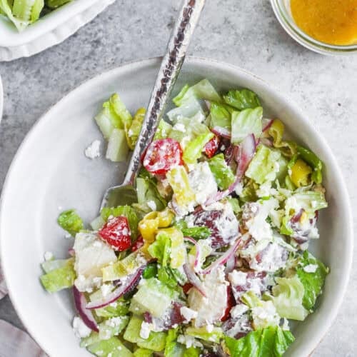 A Bowl Of Fresh Tarpon Springs Greek Salad With Potato Salad, Lettuce, Tomatoes, Cucumber, Red Onion, Feta Cheese, And Dressing On The Side, Served With A Fork On A Light Gray Surface.
