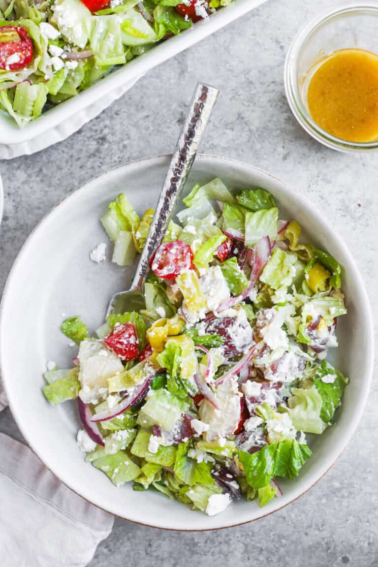 A Bowl Of Fresh Tarpon Springs Greek Salad With Potato Salad, Lettuce, Tomatoes, Cucumber, Red Onion, Feta Cheese, And Dressing On The Side, Served With A Fork On A Light Gray Surface.