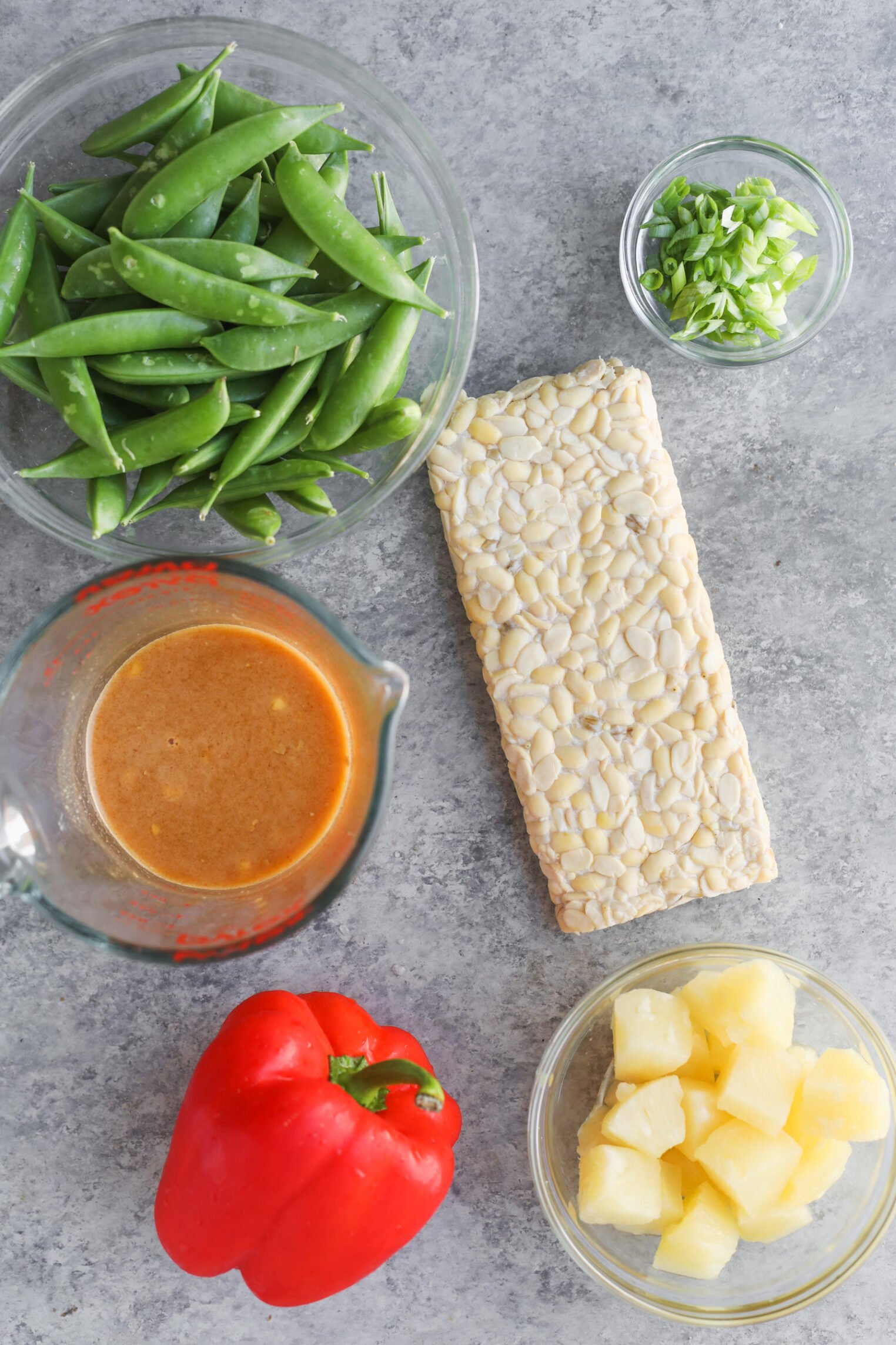 Teriyaki Tempeh Stir Fry 2 A Top-Down View Of Ingredients For Tempeh Stir Fry On A Gray Surface: Snap Peas In A Bowl, A Block Of Tempeh, Sliced Green Onions, Pineapple Chunks In A Glass Bowl, A Red Bell Pepper, And A Measuring Cup With Sauce.