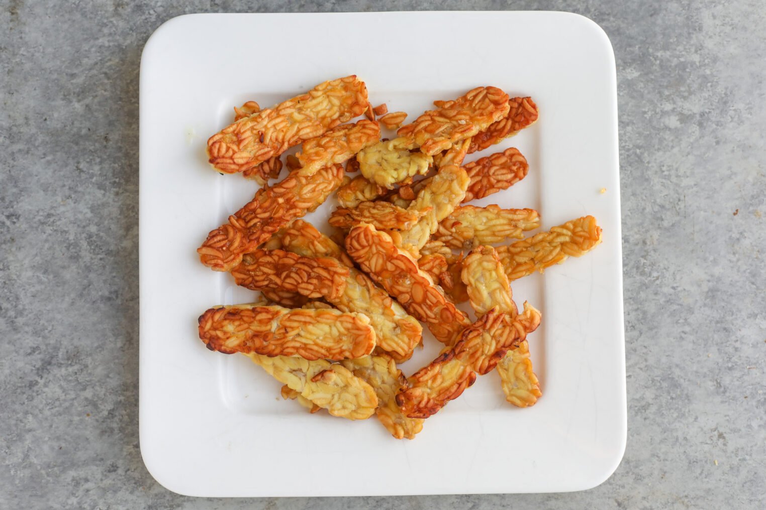 Teriyaki Tempeh Stir Fry 4 Sliced And Cooked Tempeh Stir Fry Pieces Arranged On A Square White Plate, Placed On A Gray Textured Surface.