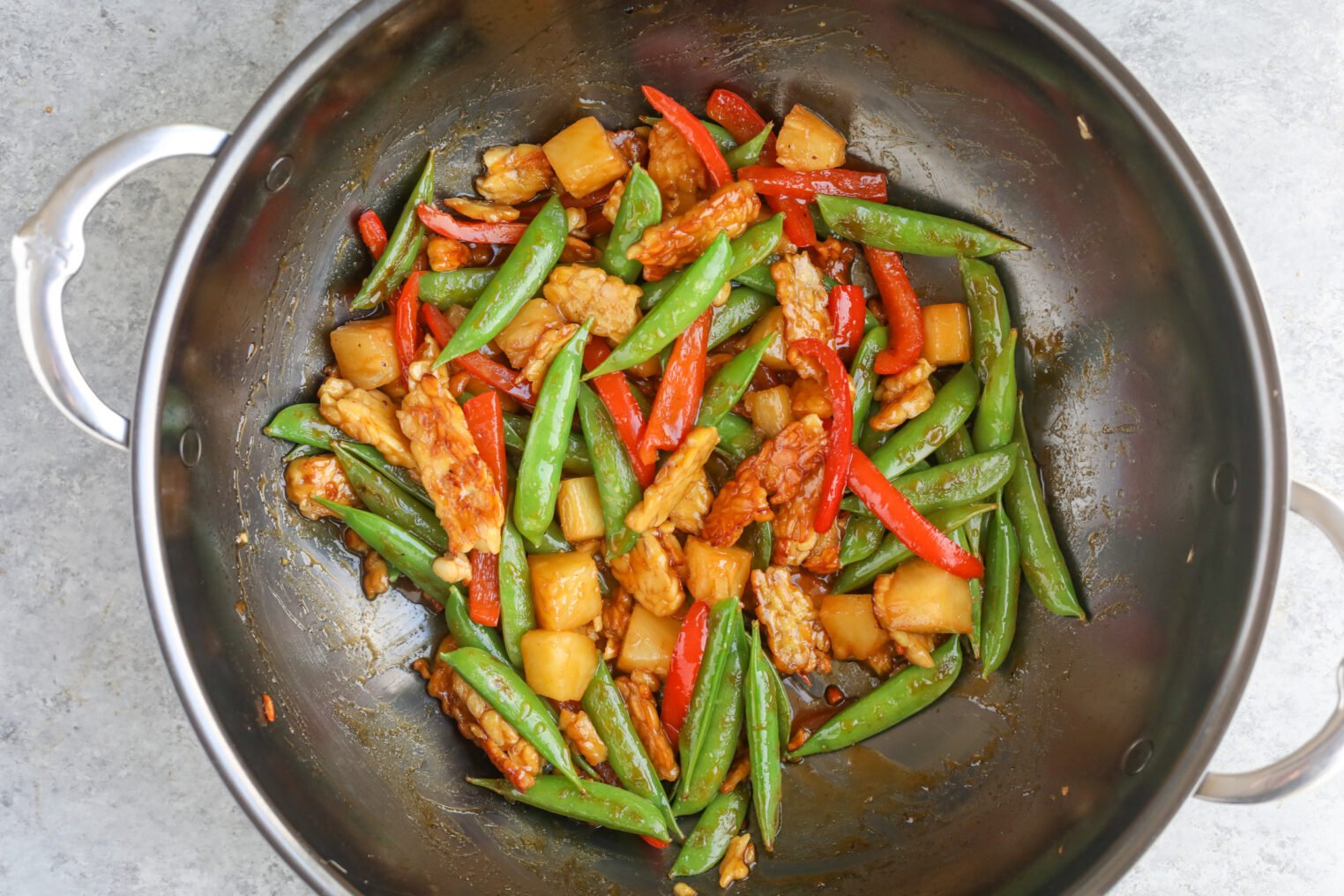 Teriyaki Tempeh Stir Fry 6 A Tempeh Stir Fry Sizzles In A Metal Wok With Snap Peas, Red Bell Pepper Strips, And Pineapple Chunks, All Coated In A Savory Brown Sauce.