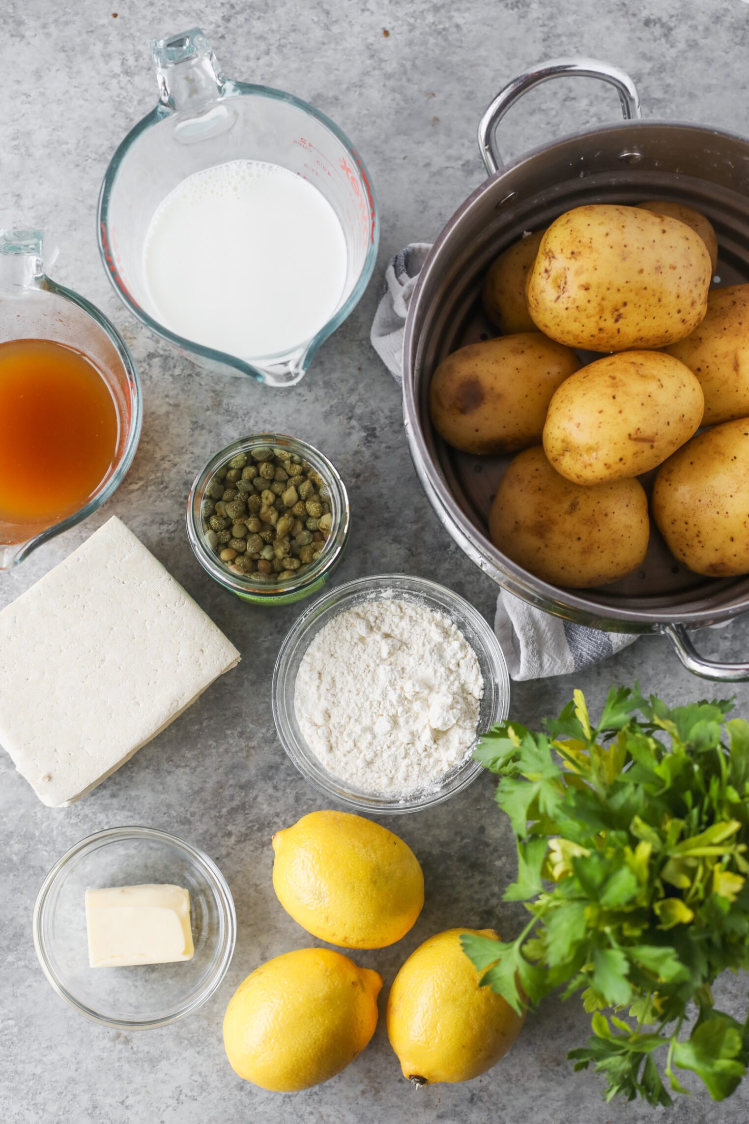 Overhead View Of Ingredients On A Gray Countertop: Whole Potatoes In A Pot, Milk, Broth, Capers, Flour, Butter, White Cheese, Three Lemons, And Fresh Parsley—Perfect For Creating A Delicious Tofu Piccata-Inspired Dish.