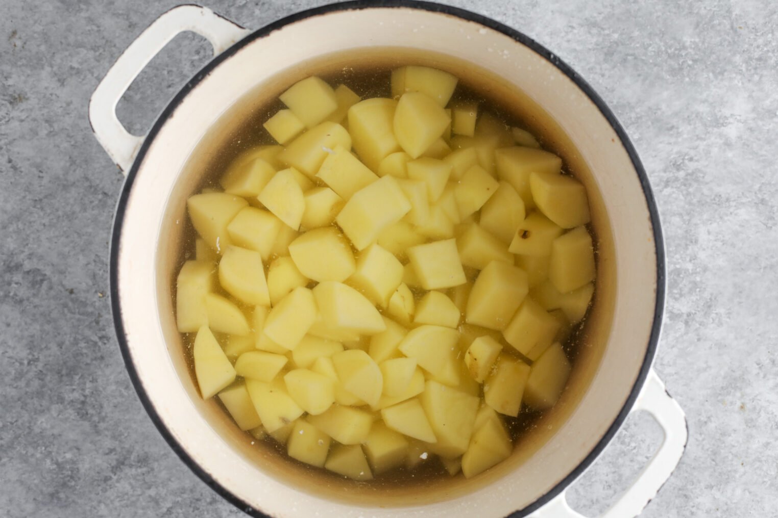 A White Pot Filled With Peeled And Chopped Potatoes Submerged In Water, Resting On A Gray Countertop—Perfect For Prepping Sides To Pair With Tofu Piccata.