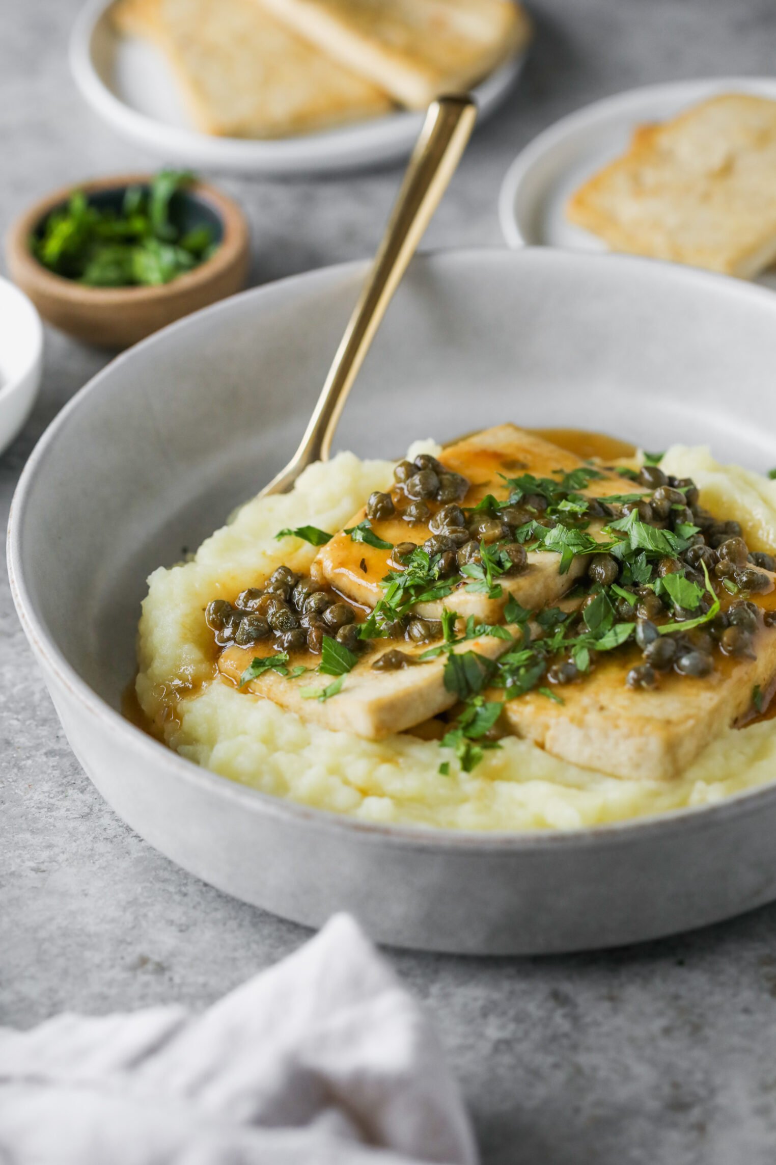 A Bowl Of Mashed Potatoes Topped With Sliced Tofu Piccata, Capers, Fresh Herbs, And Brown Sauce, Served With A Gold Fork. Plates With Bread And A Small Bowl Of Chopped Herbs Are In The Background.