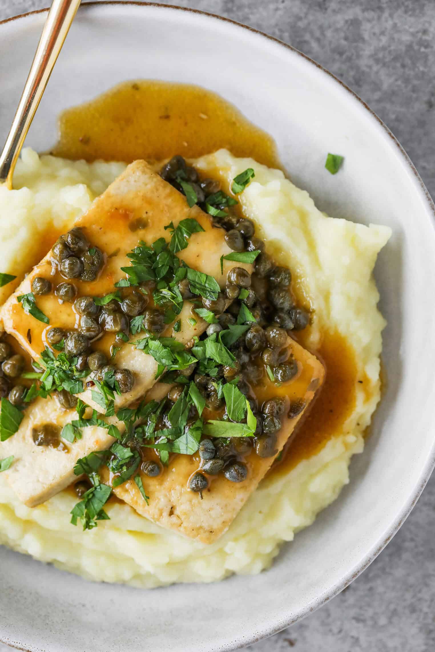 A Plate Of Mashed Potatoes Topped With Tofu Piccata, Caper Sauce, Chopped Fresh Parsley, And Brown Gravy, Served With A Gold-Handled Fork On A Light-Colored Plate.