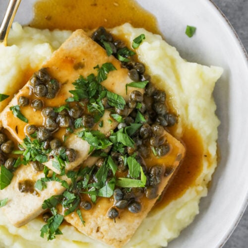 A White Plate With Creamy Mashed Potatoes Topped With A Tofu Piccata Steak, Capers, Brown Sauce, And Chopped Fresh Parsley. A Gold Fork Rests On The Edge Of The Plate.