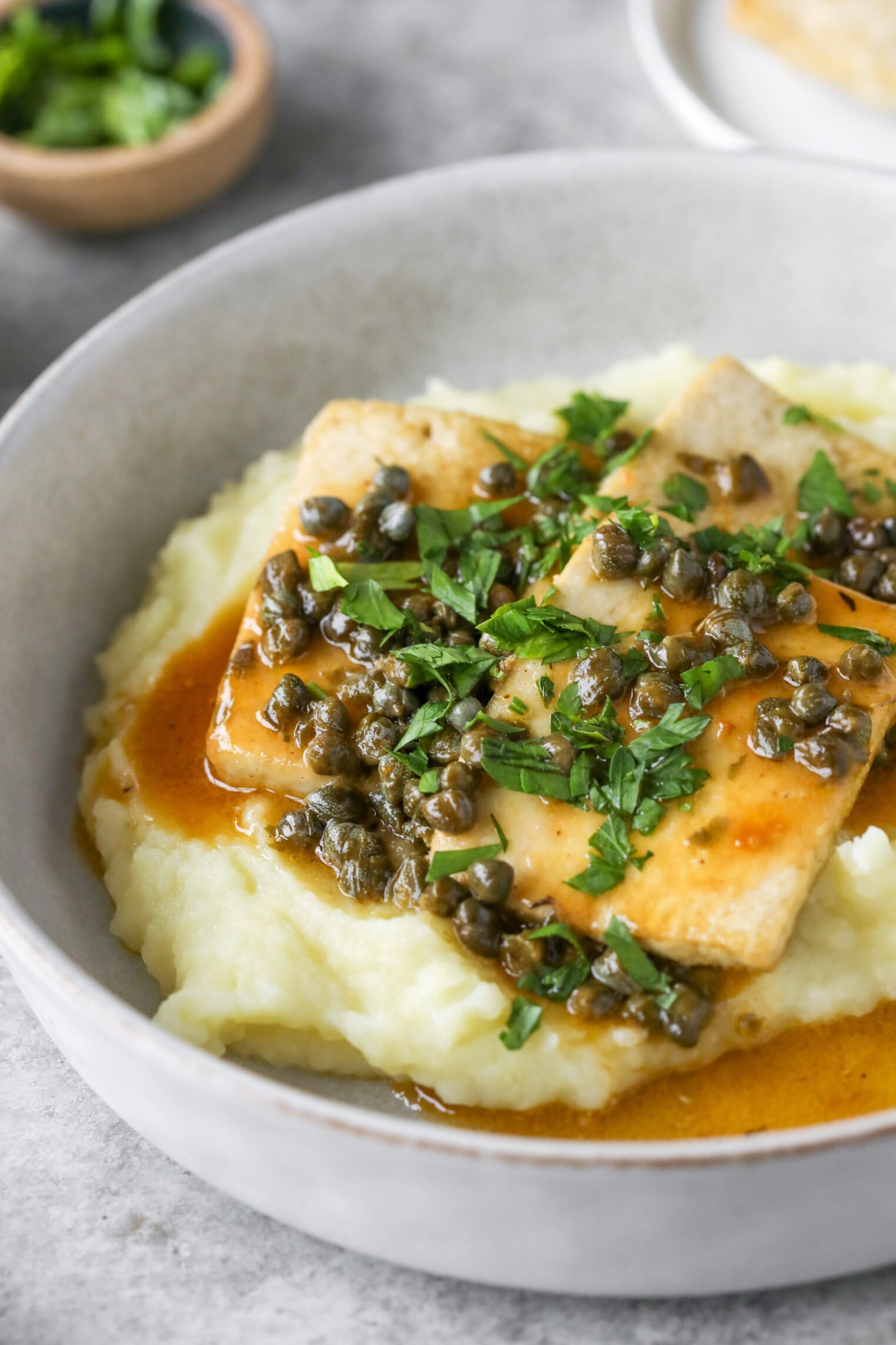 A Plate Of Mashed Potatoes Topped With Two Pieces Of Fish Fillet, Capers, Sauce, And Chopped Fresh Parsley Evokes The Flavors Of Classic Tofu Piccata. A Small Bowl Of Herbs Is Blurred In The Background.