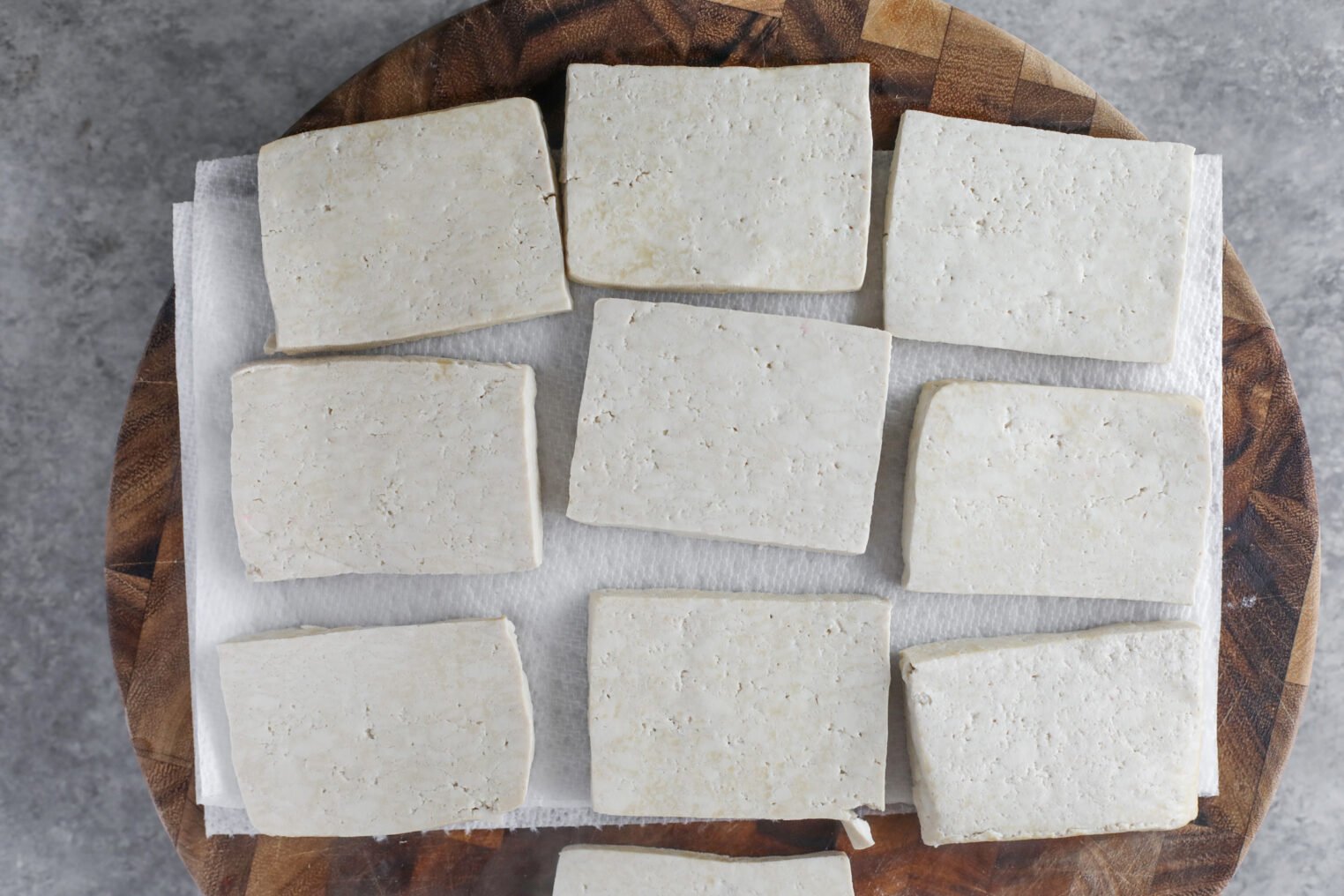 Rectangular Slices Of Tofu, Soon To Become Tofu Piccata, Are Arranged In Rows On A Paper Towel Atop A Wooden Cutting Board, Viewed From Above.
