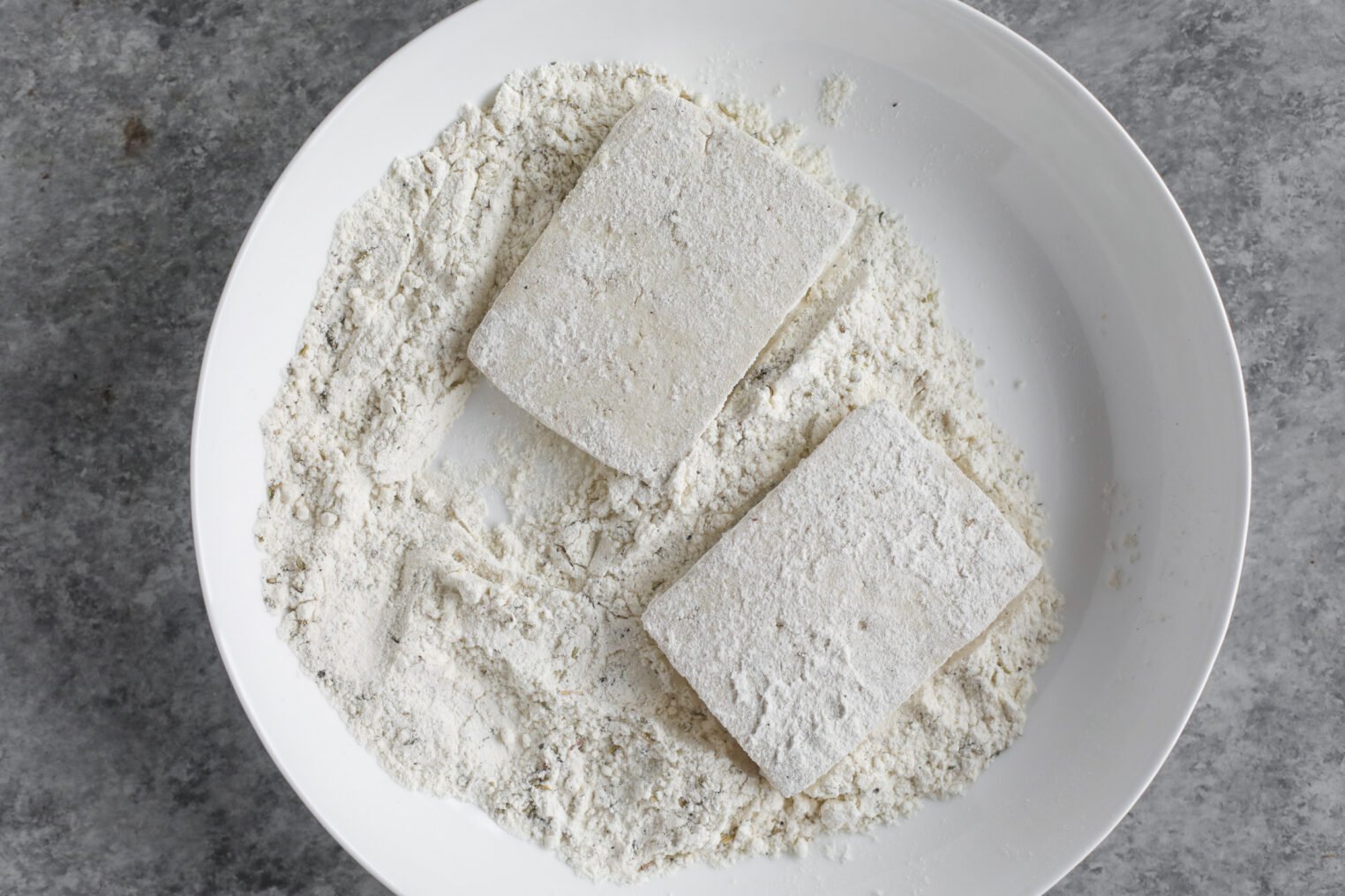 Two Rectangular Pieces Of Tofu, Perfect For Tofu Piccata, Are Coated In Flour And Rest On A White Plate Filled With Flour, Set Atop A Gray Surface.
