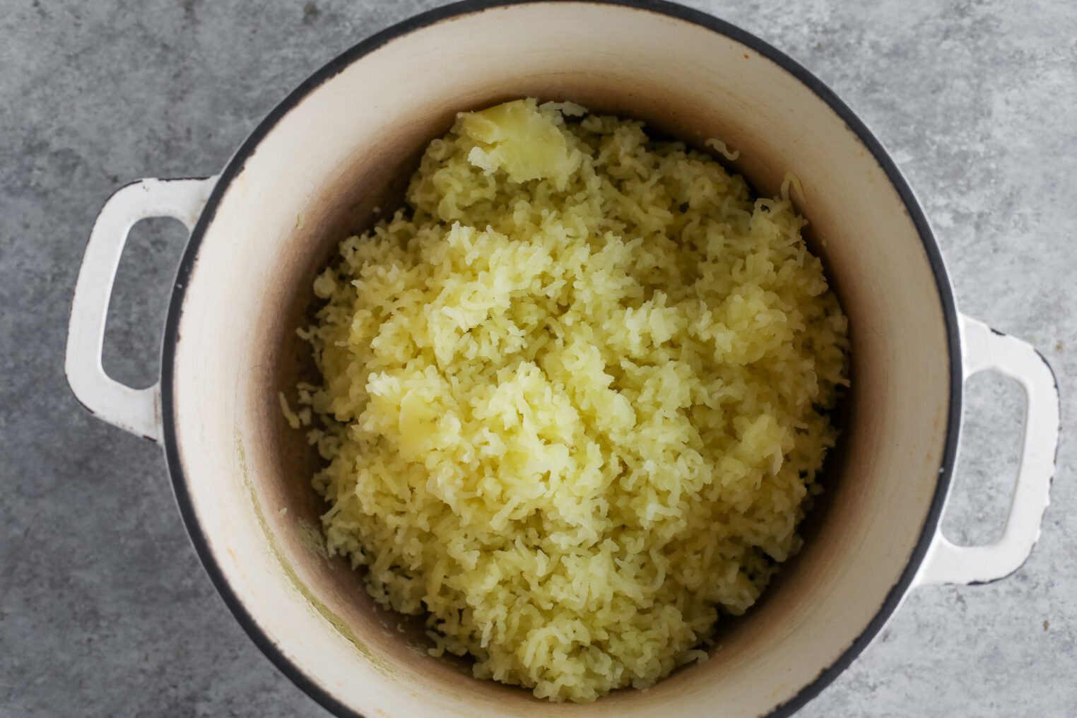 A Top View Of Cooked Yellow Rice With Chunks Of Potato And Tofu Piccata In A White Pot On A Gray Surface.