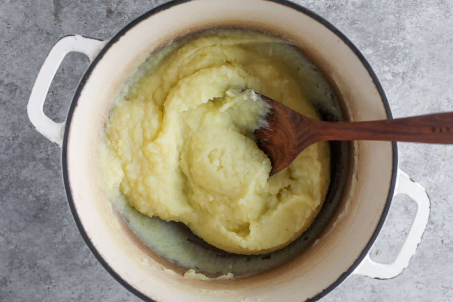 A Pot Of Mashed Potatoes Being Stirred With A Wooden Spoon, Shown From Above On A Gray Surface—Perfect As A Side For Tofu Piccata.