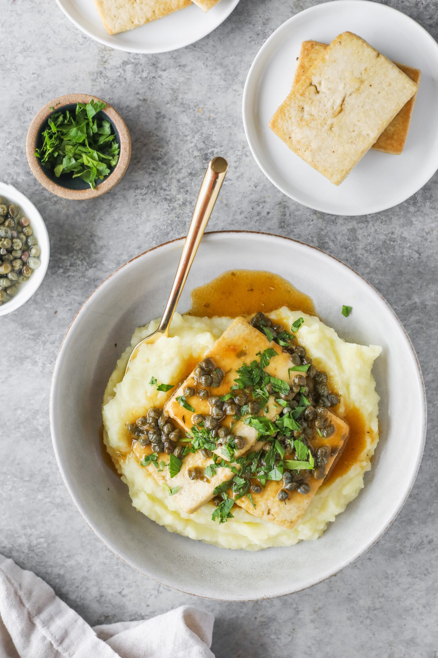 A Bowl Of Mashed Potatoes Topped With Tofu Piccata, Capers, Sauce, And Chopped Herbs. Nearby Are Small Plates With Extra Tofu, A Bowl Of Chopped Herbs, And A Bowl Of Capers On A Gray Surface. A Fork Rests On The Bowl.
