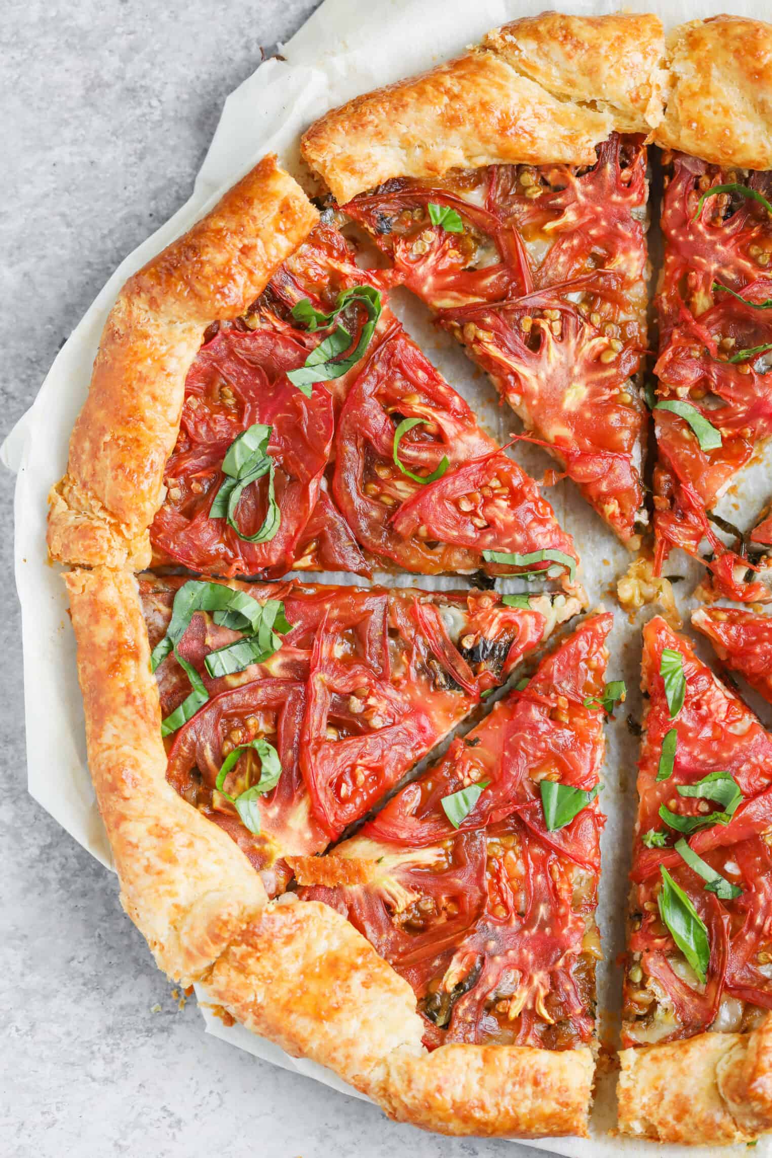 Tomato Galette 1 A Rustic Tomato Galette With A Golden, Flaky Crust Is Sliced Into Pieces. Fresh Tomato Slices And Basil Leaves Crown The Savory Tomato Galette, Which Rests On Parchment Paper Atop A Gray Surface.