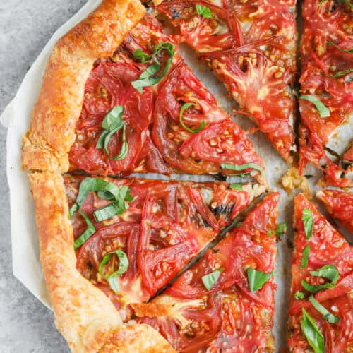 Tomato Galette 14 A Rustic Tomato Galette With A Golden, Flaky Crust Is Sliced Into Pieces. Fresh Tomato Slices And Basil Leaves Crown The Savory Tomato Galette, Which Rests On Parchment Paper Atop A Gray Surface.