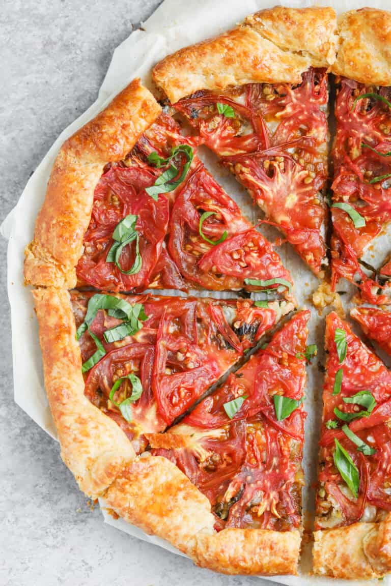A Rustic Tomato Galette With A Golden, Flaky Crust Is Sliced Into Pieces. Fresh Tomato Slices And Basil Leaves Crown The Savory Tomato Galette, Which Rests On Parchment Paper Atop A Gray Surface.