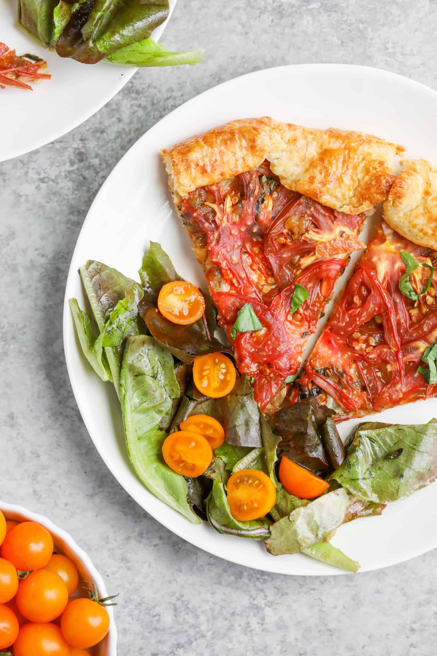 Tomato Galette 2 A White Plate With Two Slices Of Tomato Galette And A Green Salad Topped With Halved Yellow Cherry Tomatoes. Nearby Sit A Bowl Of Whole Yellow Cherry Tomatoes And Part Of Another Plate Featuring Tomato Galette And Salad.