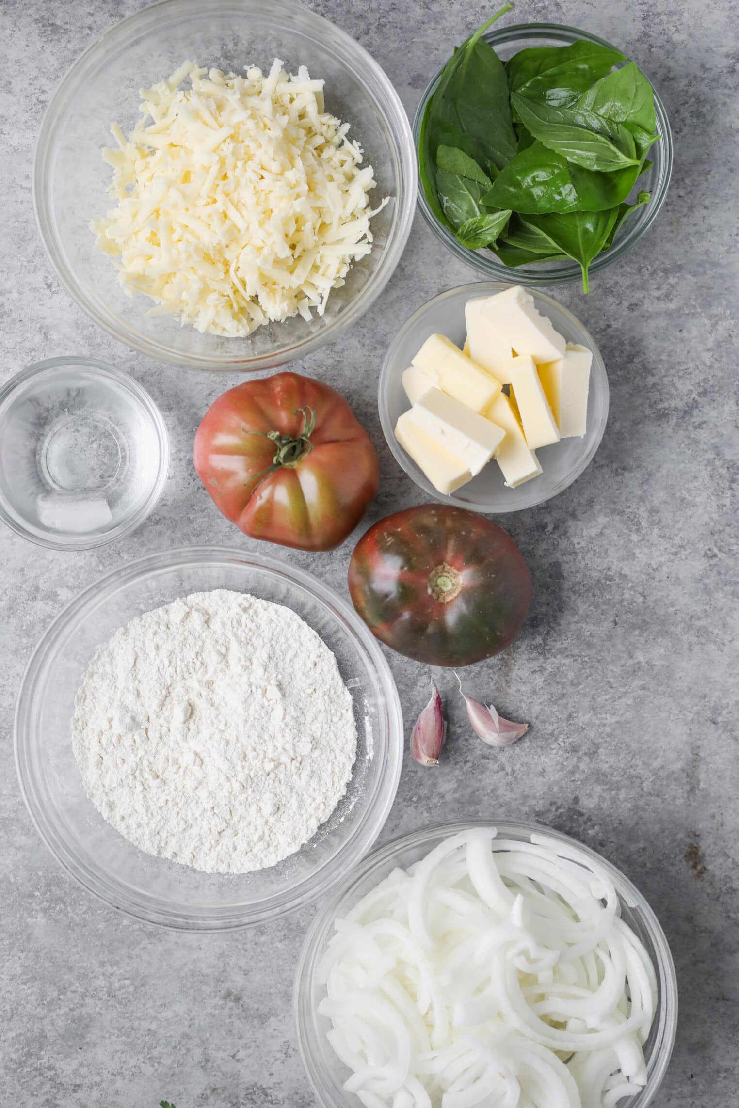 Tomato Galette 3 A Top-Down View Of Ingredients For A Tomato Galette On A Gray Surface, Including Shredded Cheese, Fresh Spinach, Two Tomatoes, Butter Cubes, Flour, Sliced Onions, Garlic Cloves, And A Small Bowl Of Water.