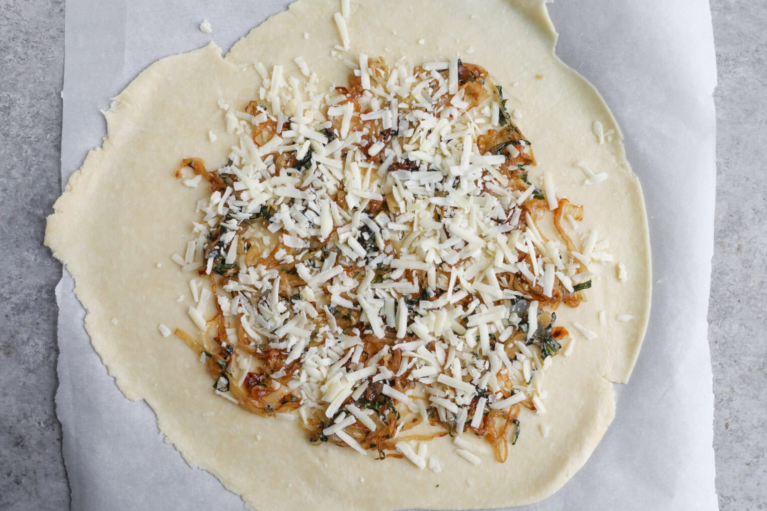 Tomato Galette 7 Unbaked Round Pastry Dough, Like A Tomato Galette, Is Topped With Cooked Onions, Herbs, And Shredded Cheese, Arranged On Parchment Paper Over A Gray Countertop.