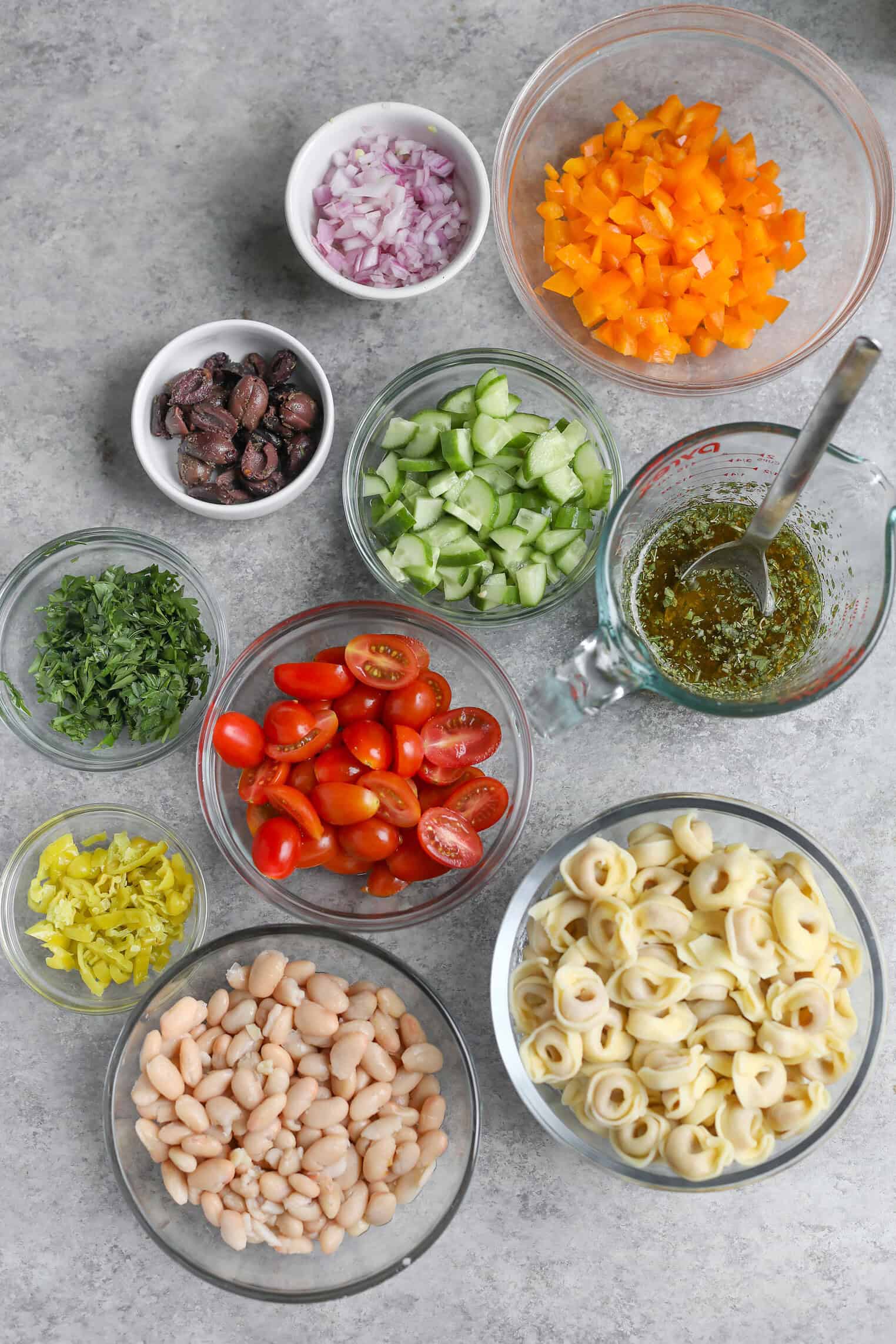 Tortellini Pasta Salad 4 Bowls Of Chopped Yellow Bell Pepper, Red Onion, Kalamata Olives, Cucumbers, Grape Tomatoes, Cooked Tortellini Pasta Salad, Cannellini Beans, Pepperoncini, Fresh Herbs, And A Glass Measuring Cup Of Dressing On A Gray Surface.