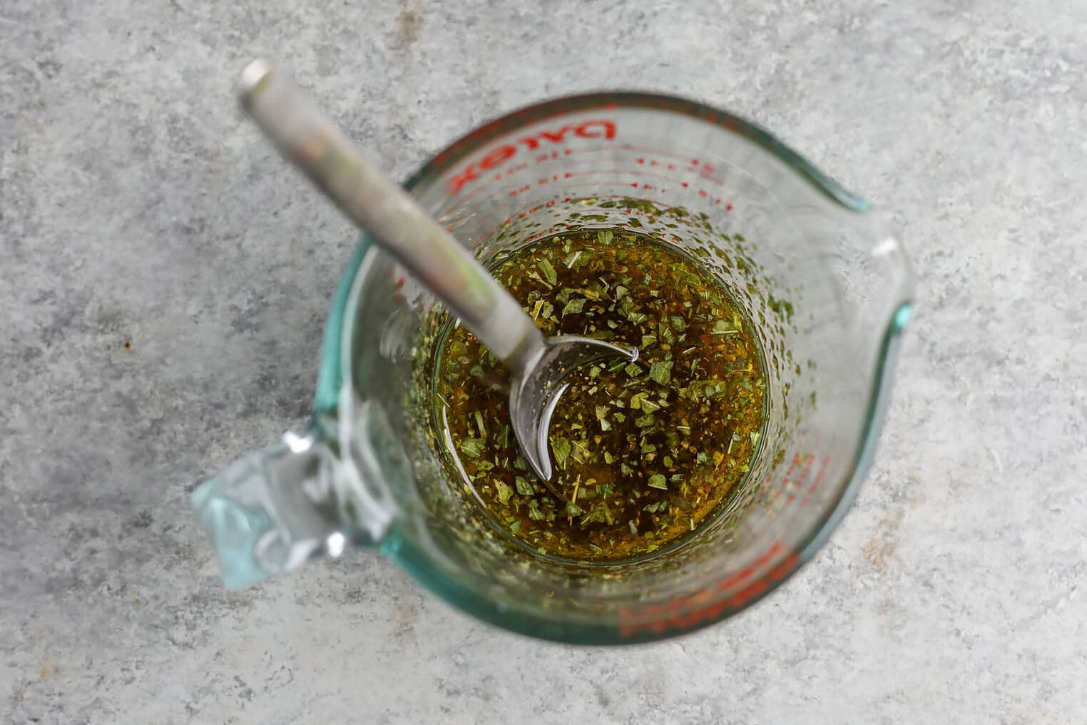 Tortellini Pasta Salad 5 A Glass Measuring Cup With A Silver Spoon Inside Contains A Herb And Oil Mixture For Tortellini Pasta Salad, Sitting On A Gray Textured Surface.