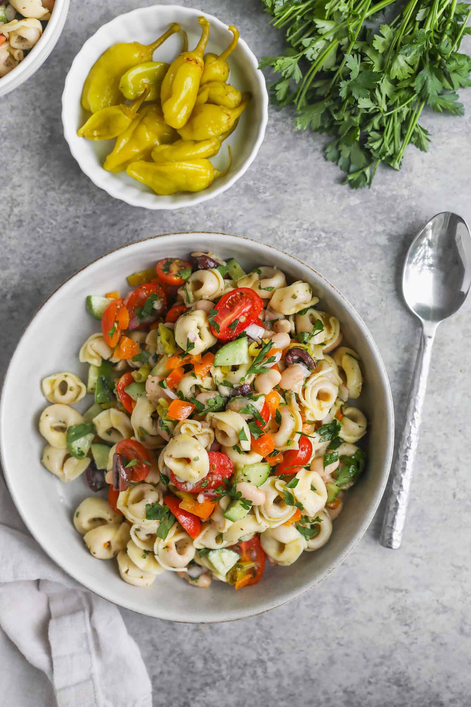 Tortellini Pasta Salad 9 A Vibrant Bowl Of Tortellini Pasta Salad With Tomatoes, Peppers, Olives, And Herbs Sits On A Gray Surface Beside A Small Bowl Of Yellow Peppers, Fresh Parsley, And A Silver Spoon.